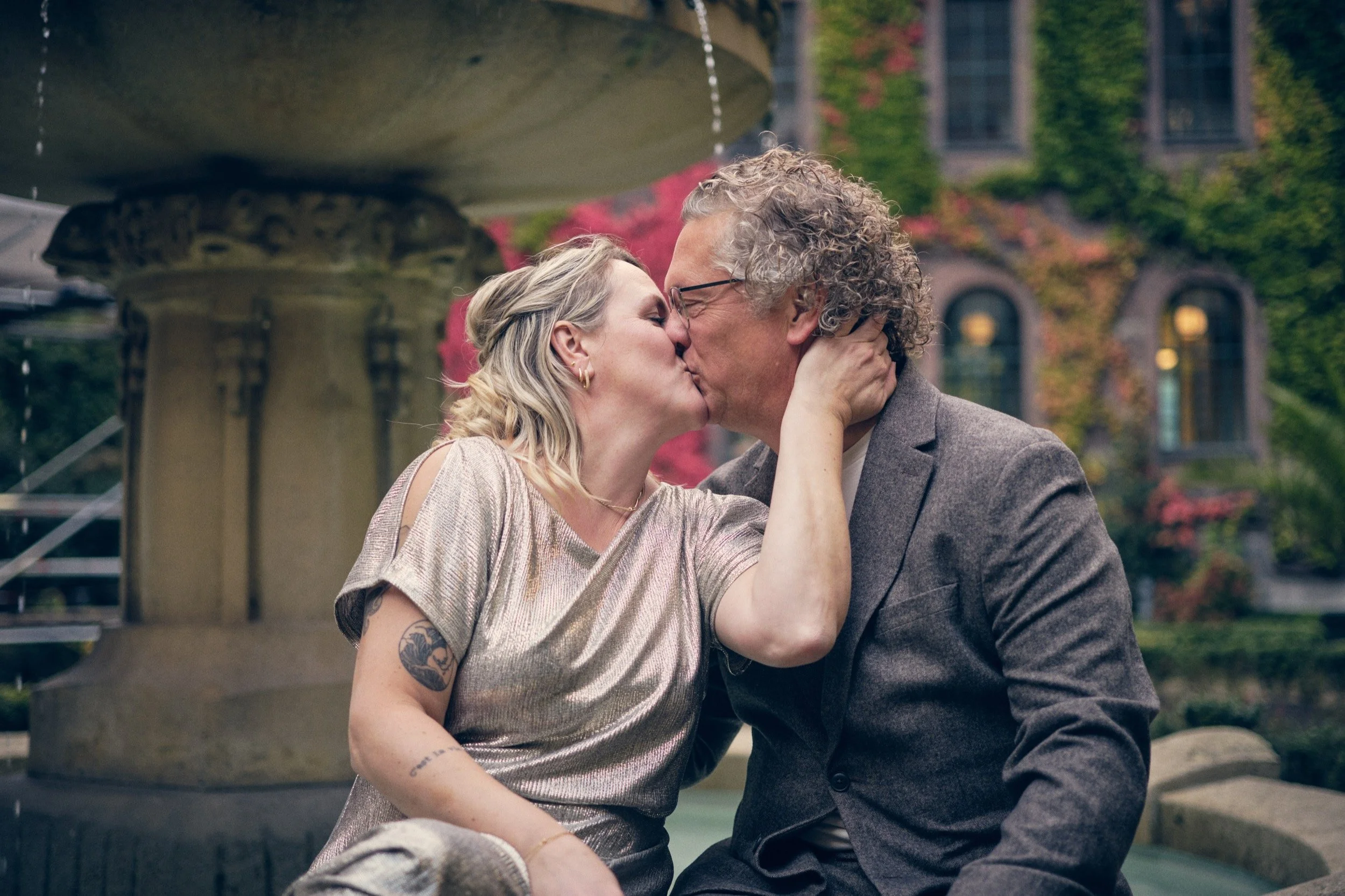 Een koppel kust elkaar op een pleintje met een fontein en een erfgoedgebouw met klimop op de achtergrond.