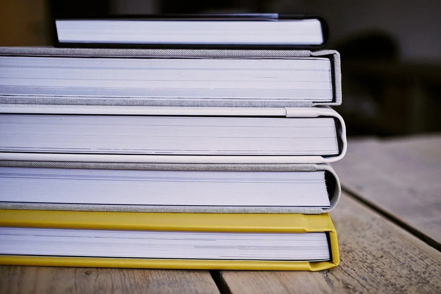 Een stapel boeken op een houten tafel, met verschillende kleuren omslagen, op de voorgrond een geel boek, en op de achtergrond een zwarte en een grijze boek.