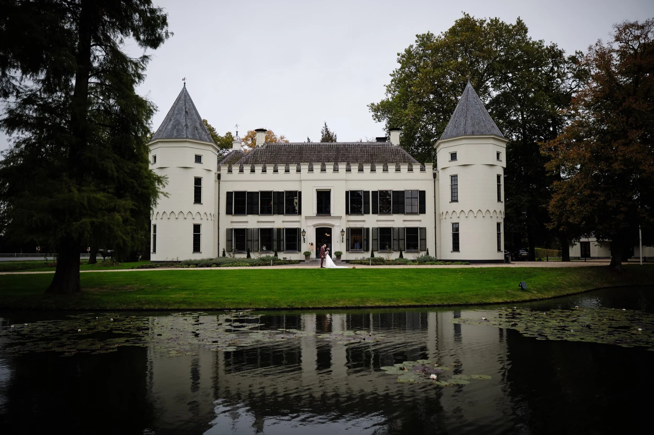 Een kasteelachtig witte woning met twee ronde torens en een paar grote bomen op de voorgrond, een grote tuin met gras en een sloot met waterlelies. Een bruid en bruidegom staan voor de ingang van het huis.