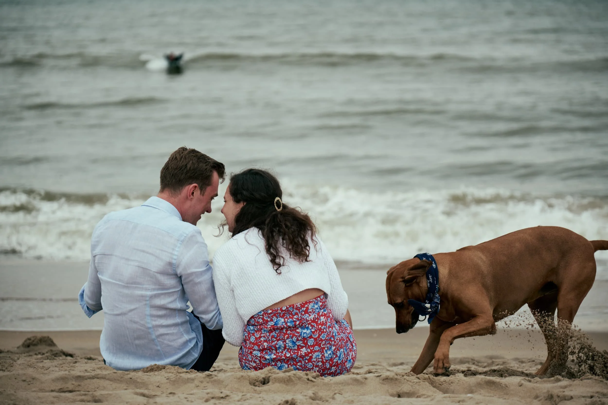 Een koppel zit knus op het strand terwijl een hond speelt in het zand, met de oceaan op de achtergrond.