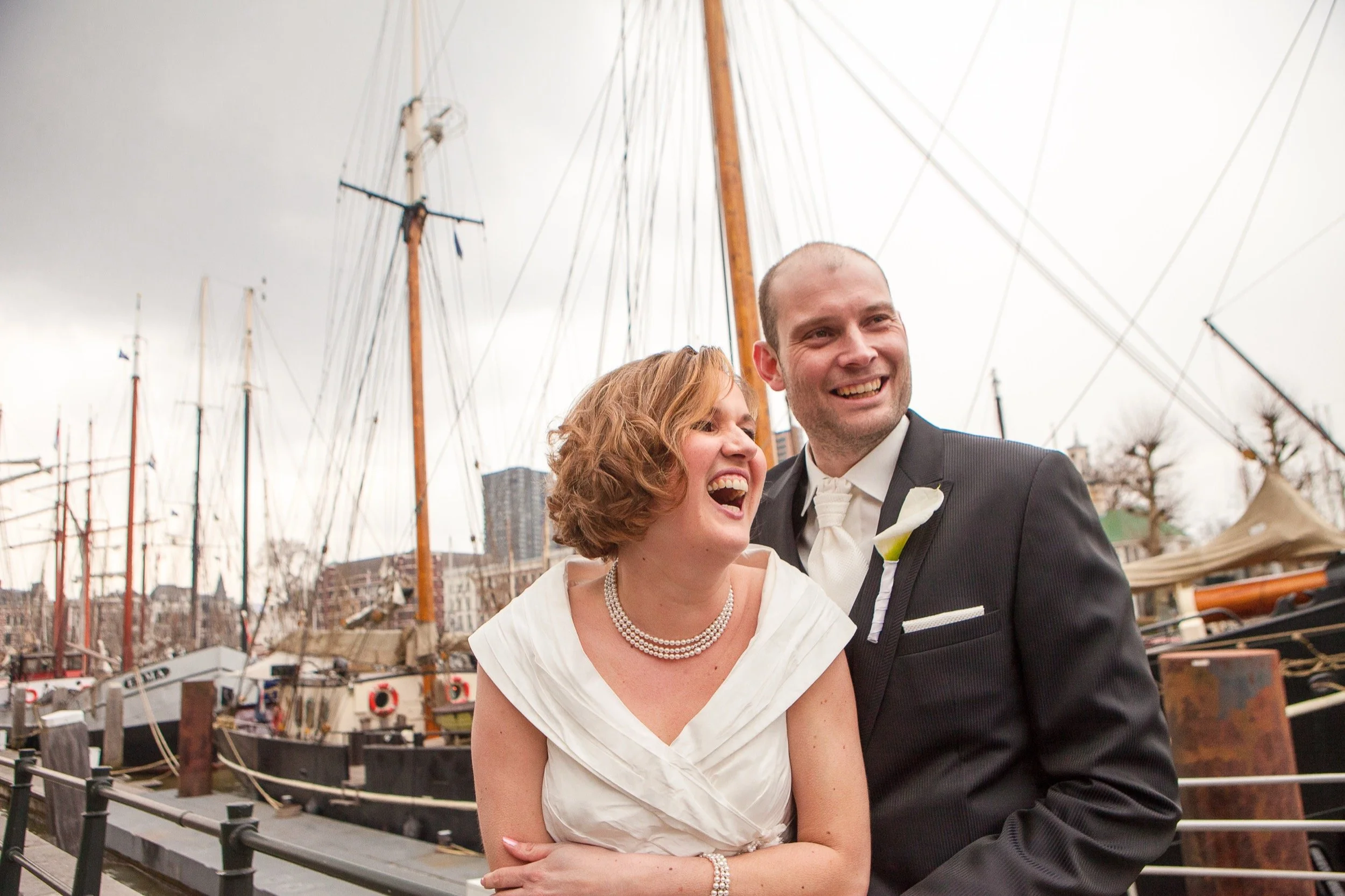 Een bruid en bruidegom lachen samen voor een haven met grote zeilboten.