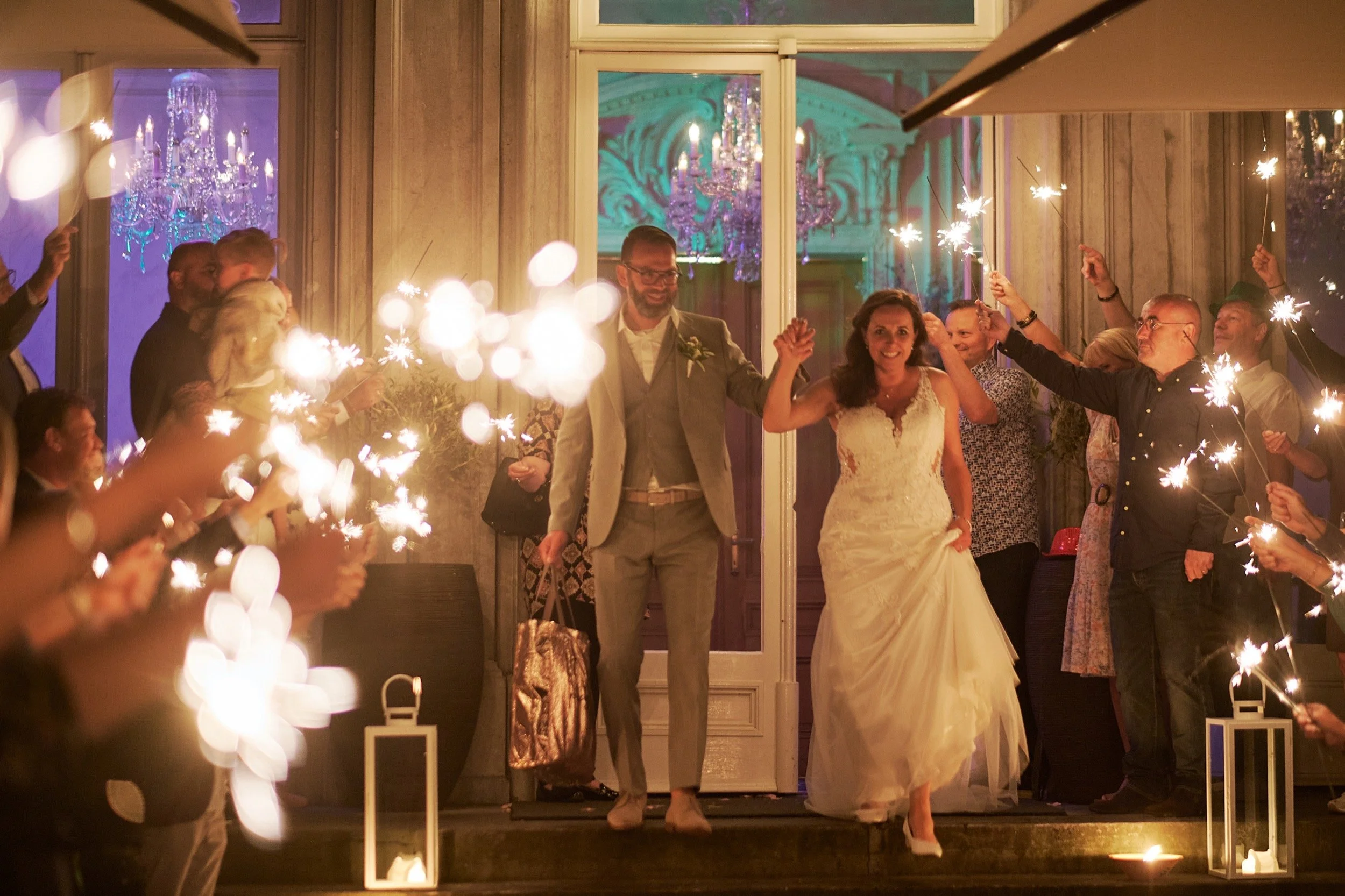 Een bruid en bruidegom die samen naar buiten lopen terwijl ze omringd worden door gasten die fakkels vasthouden. De gasten vieren de bruiloft met vuurwerk en dansen, en er zijn mooie verlichting en chandeliers zichtbaar.