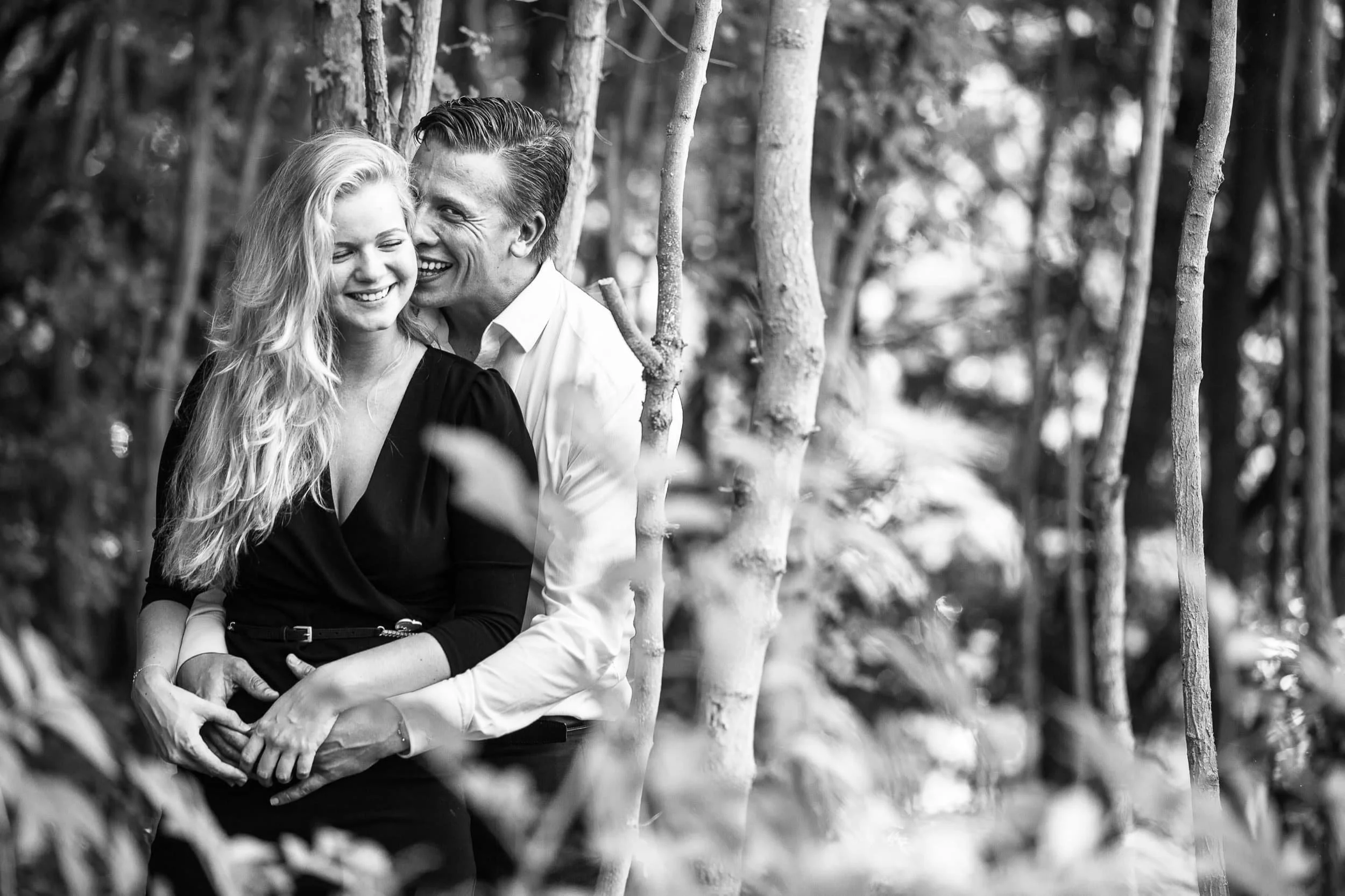 Een zwart-wit foto van een lachend paar in een bos, een man en een vrouw die dicht bij elkaar staan en elkaar vasthouden.