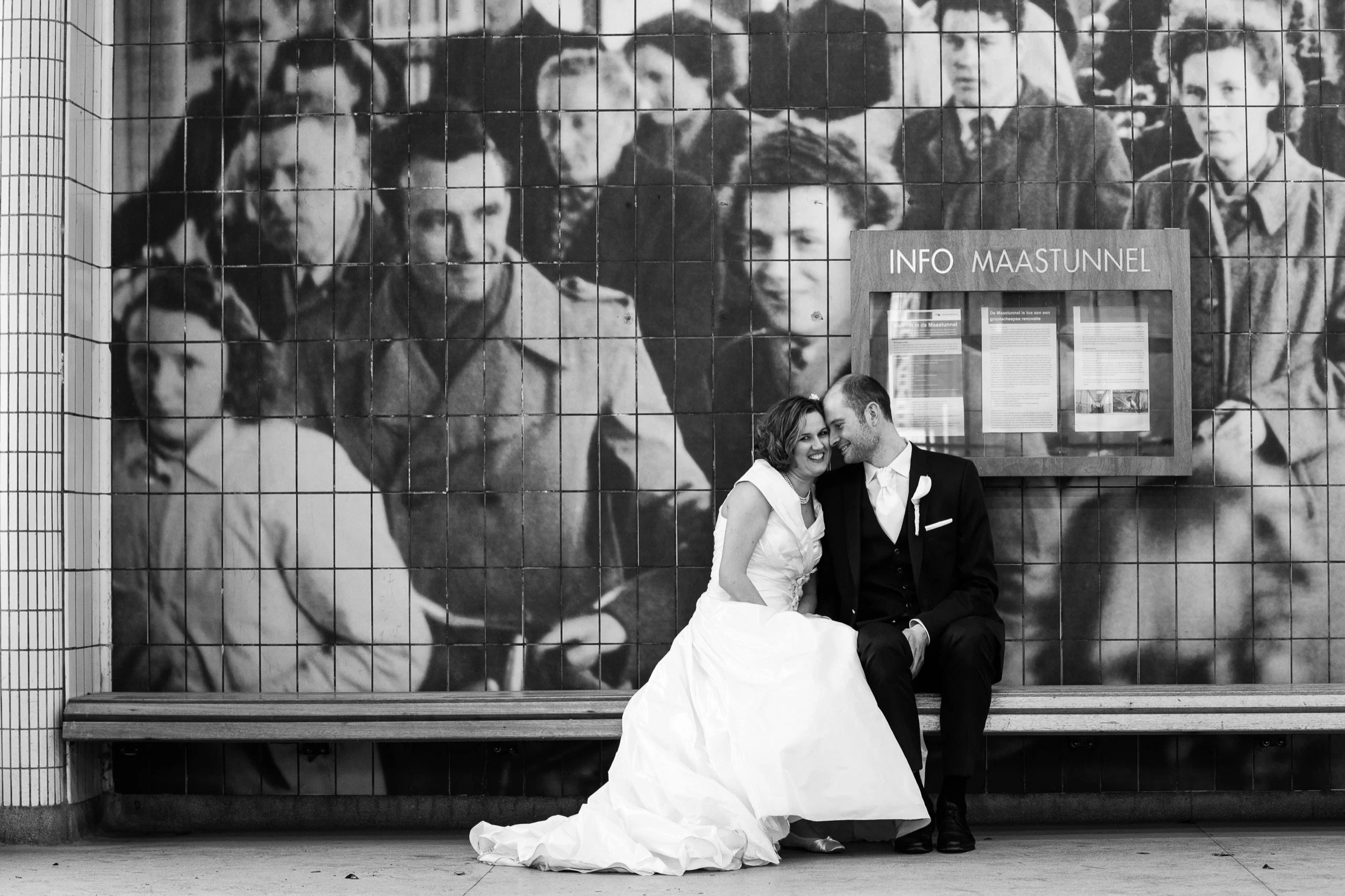 Een getrouwd stel zit samen op een bankje voor een groot zwart-wit portretfoto van mensen in een opslag of informatiehoek en lacht naar elkaar.