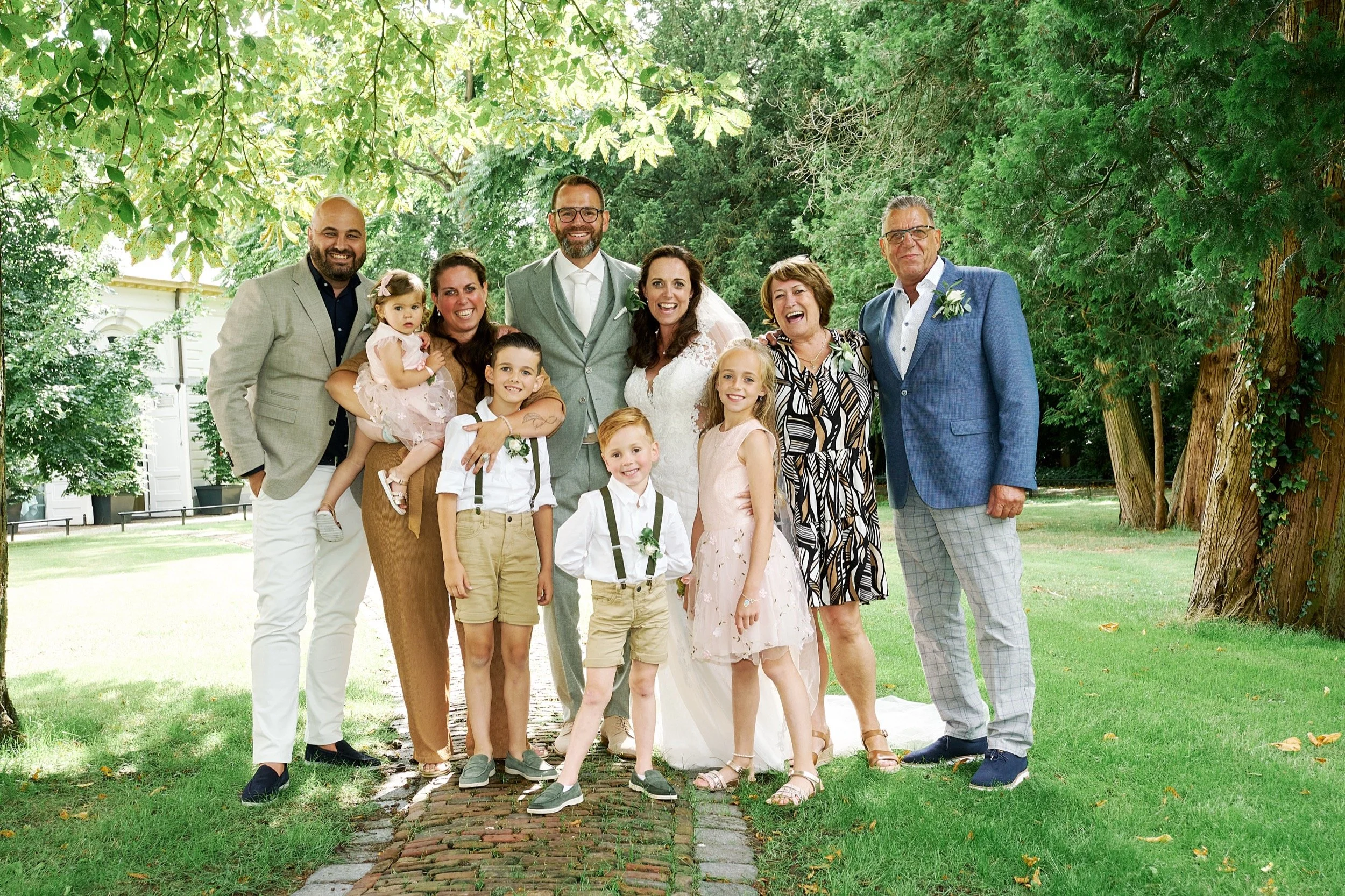 Groep mensen in een tuin tijdens een bruiloft, met een bruid en bruidegom in het midden, omringd door familie en vrienden.
