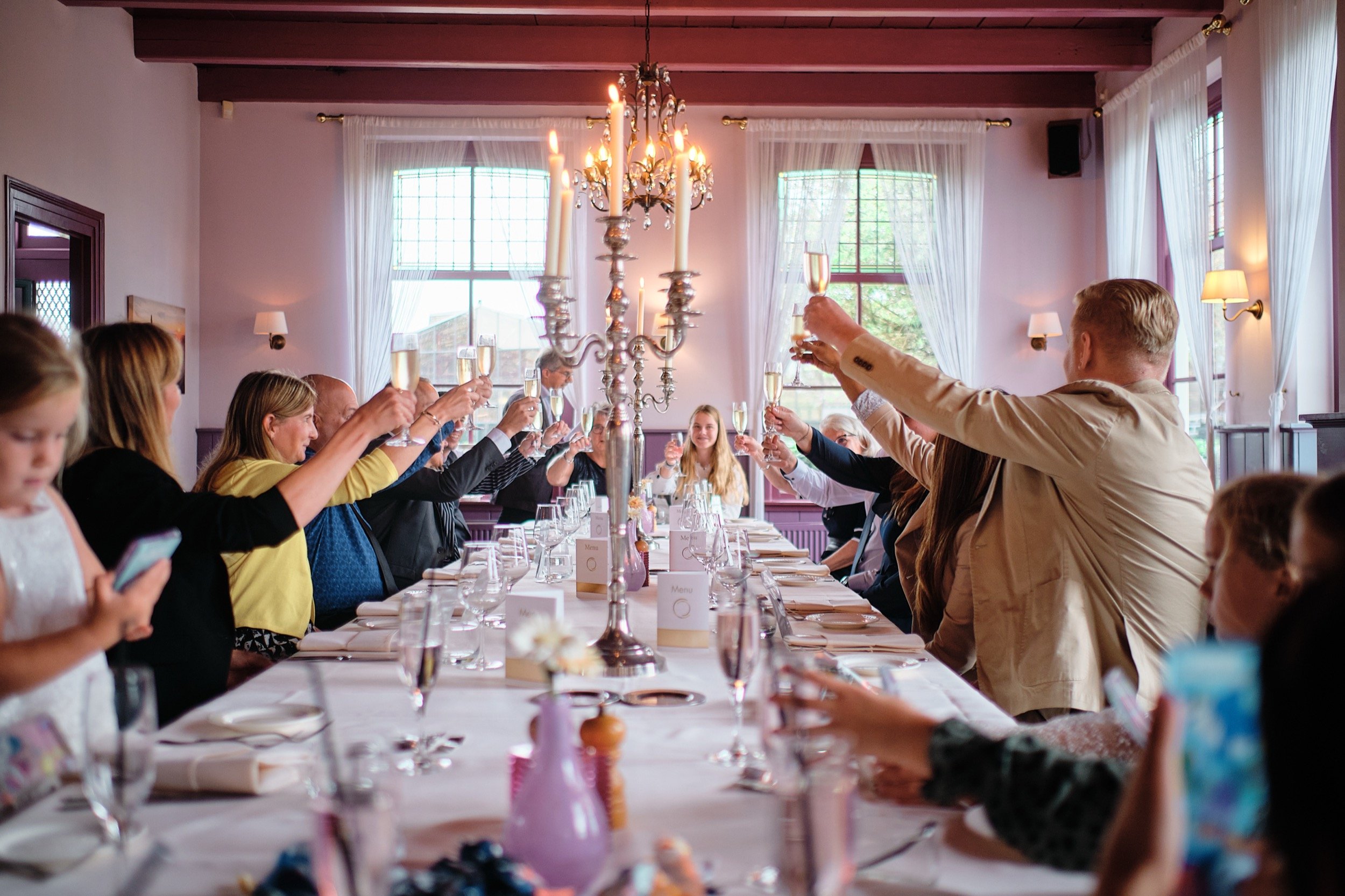 Grote groepsfoto van mensen die samen een feestje vieren, zittend aan een lange tafel, met champagneglazen omhoog in een toast, in een nette zaal met grote ramen, witte gordijnen en een kroonluchter in het midden.
