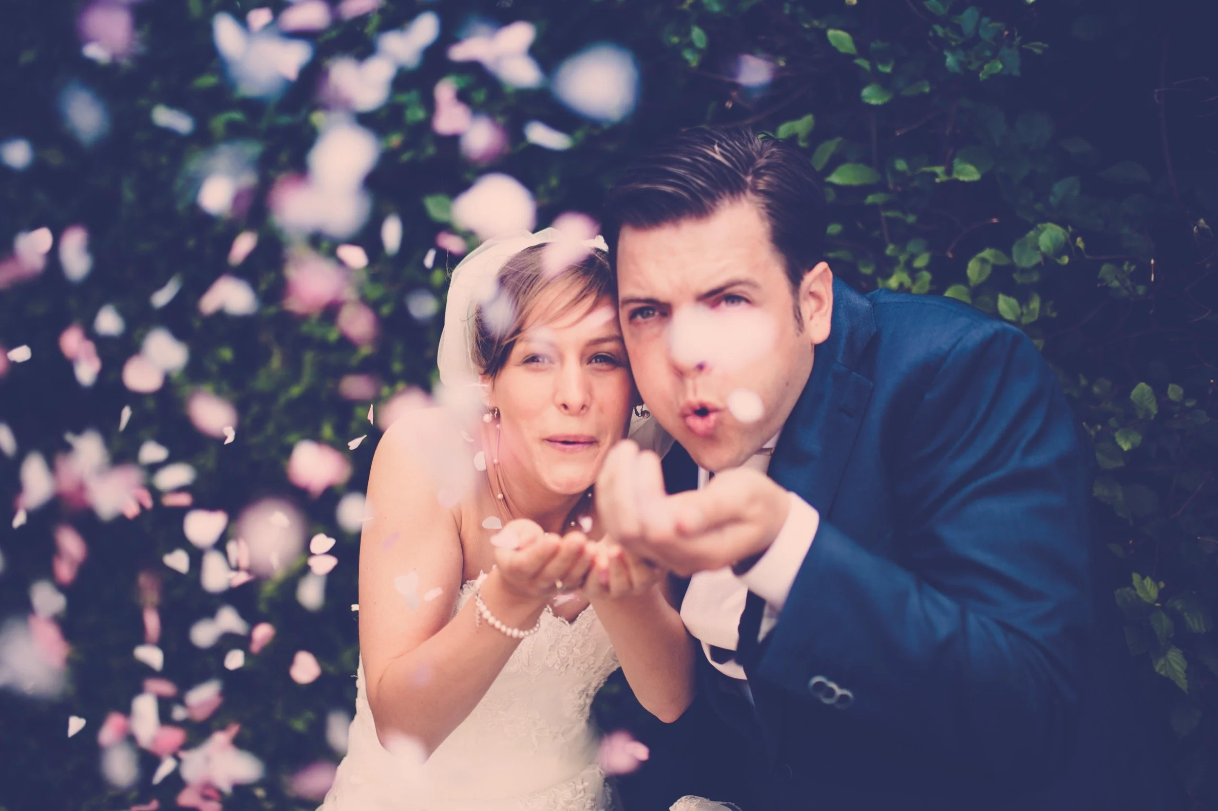 Een getrouwd stel in trouwkleding onder een bloemenboog, ze blazen confetti op.