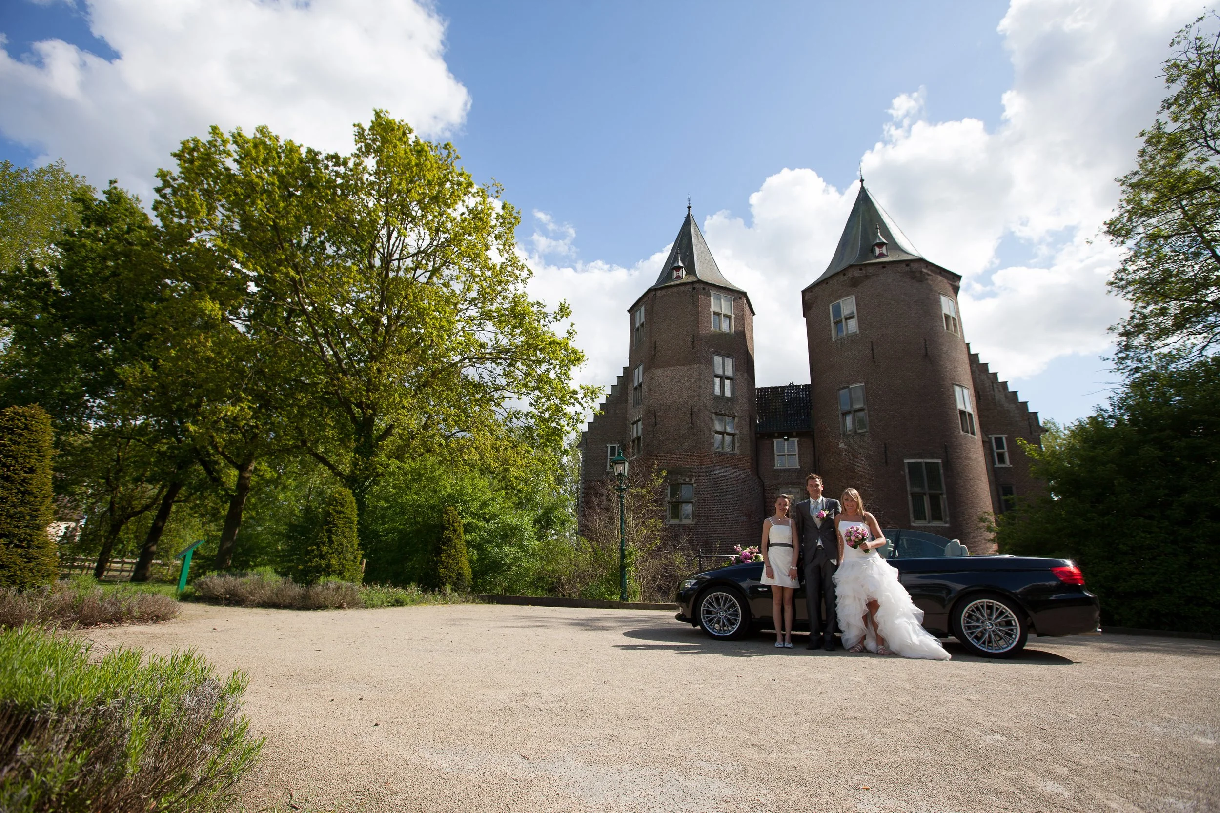 Een bruid en bruidegom staan naast een zwarte auto voor een kasteel met twee torens, omringd door groene bomen op een zonnige dag.