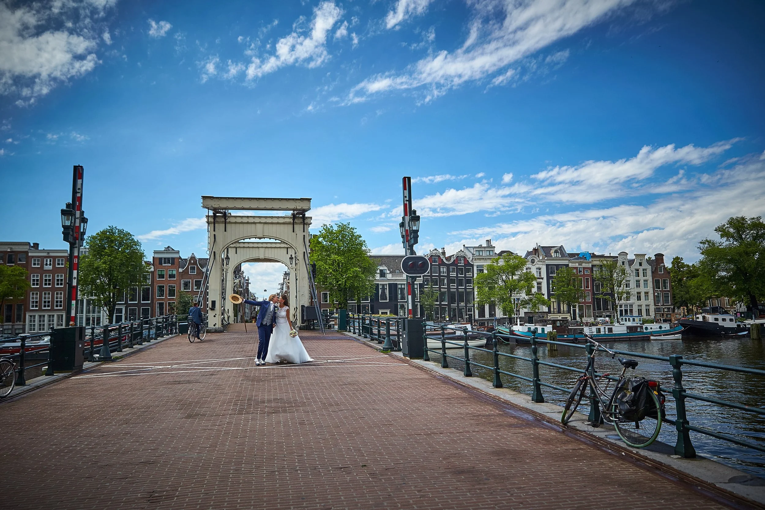 Een paar dat getrouwd is, staat op een brug in Amsterdam, met water en boten links en traditionele grachtenhuizen op de achtergrond.