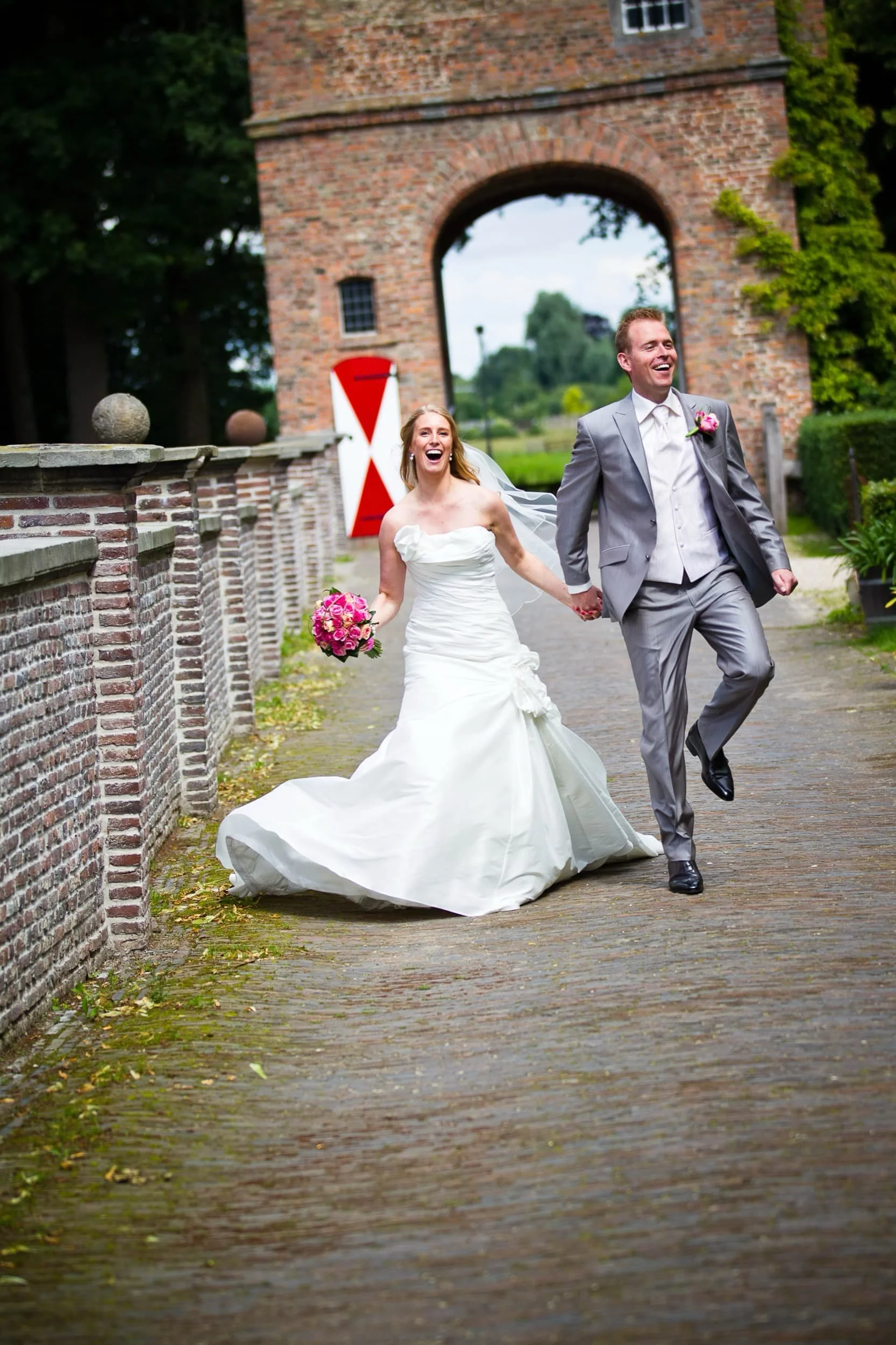 Gelukkig bruid en bruidegom lopen hand in hand, lachend, op een kasseienweg voor een oude bricken poort met een boog en een rood-wit wegwijzer in de achtergrond, tijdens een huwelijksfotografie.