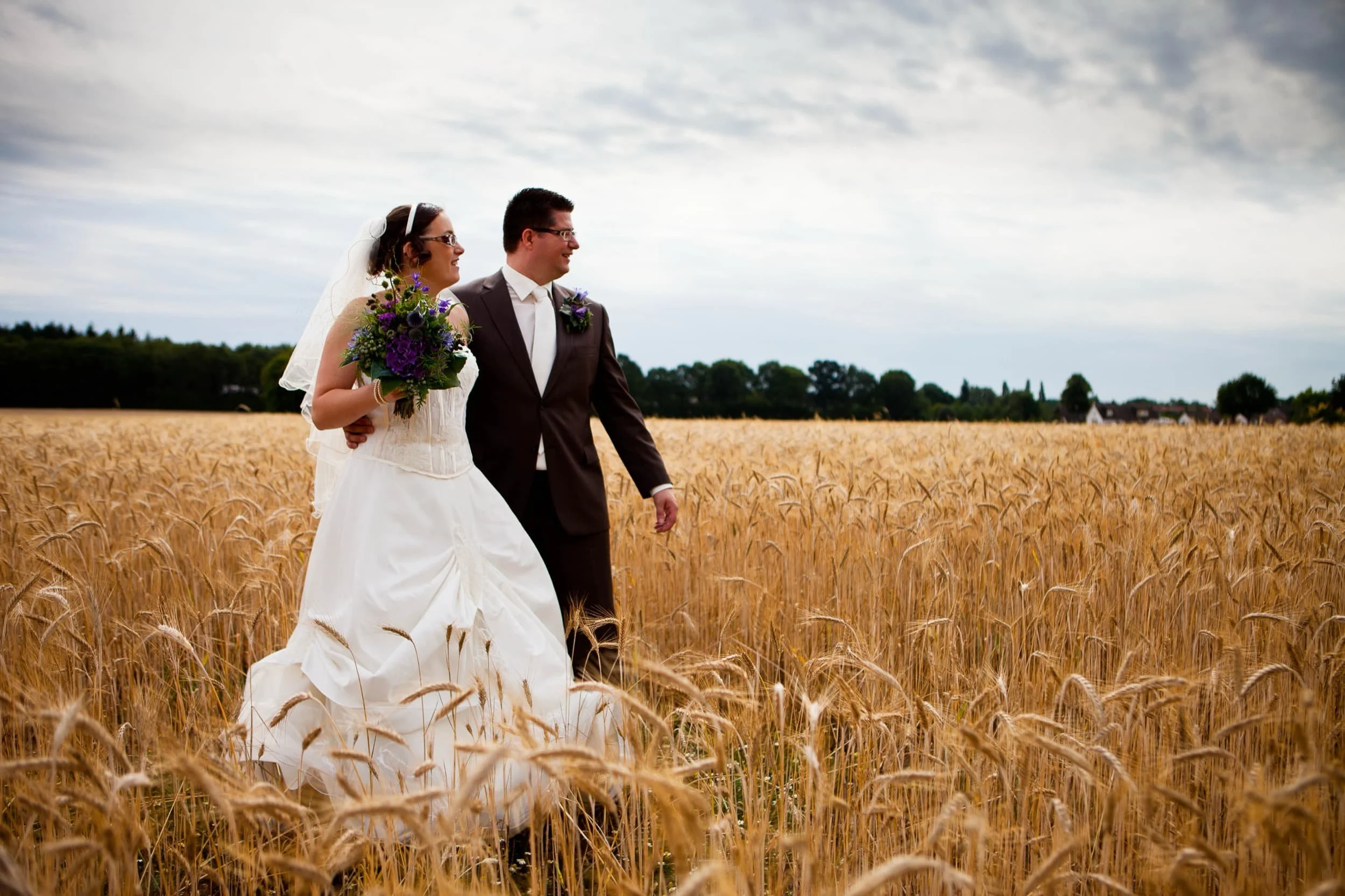 Bruidspaar wandelen door een goudgele tarweveld op hun huwelijksdag, onder een bewolkte hemel.