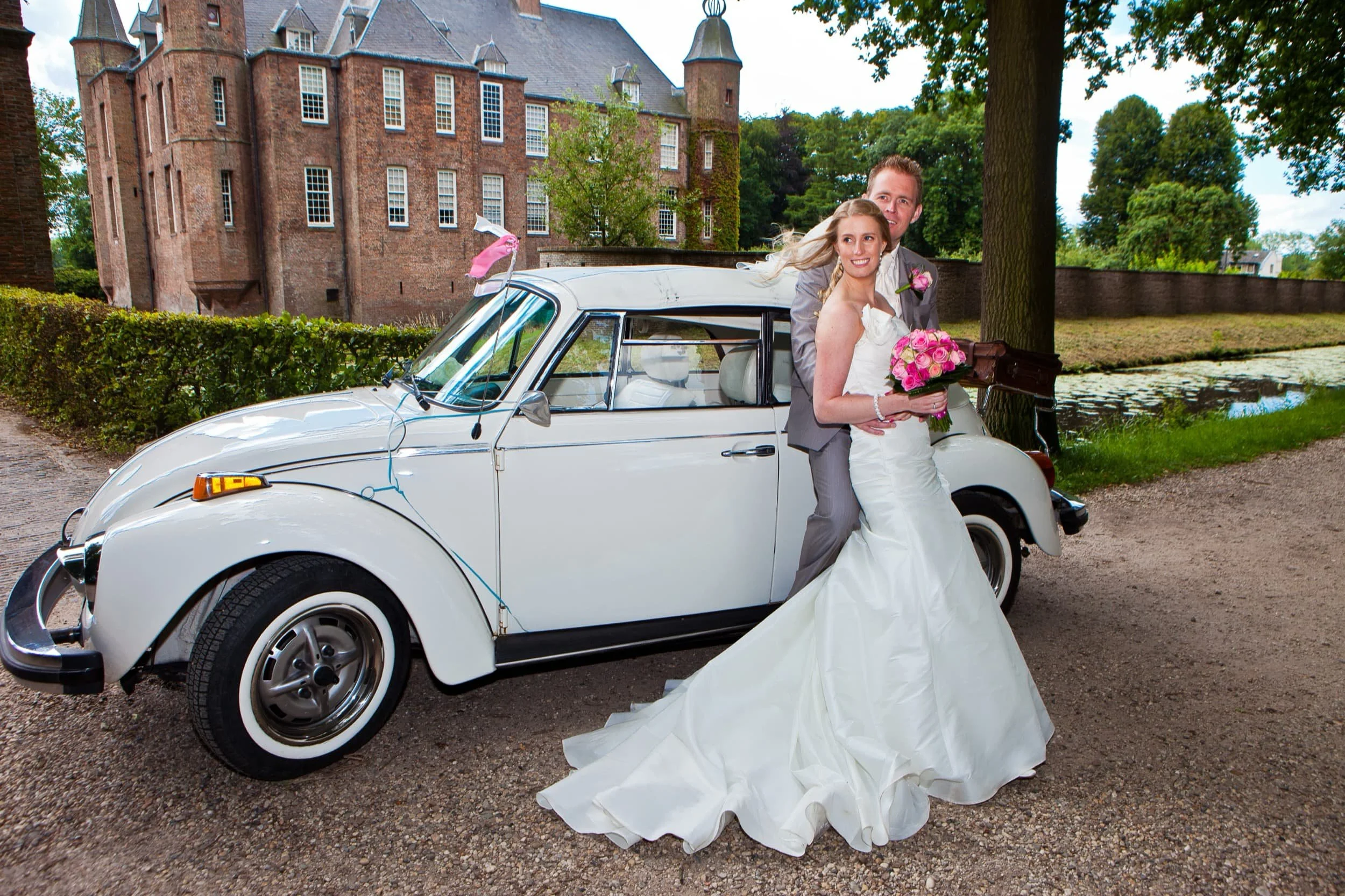 Een bruid en bruidegom in trouwkleding die naast een witte oldtimer auto staan, met een bruidss bouquet, buiten in een tuin met een kasteel op de achtergrond.
