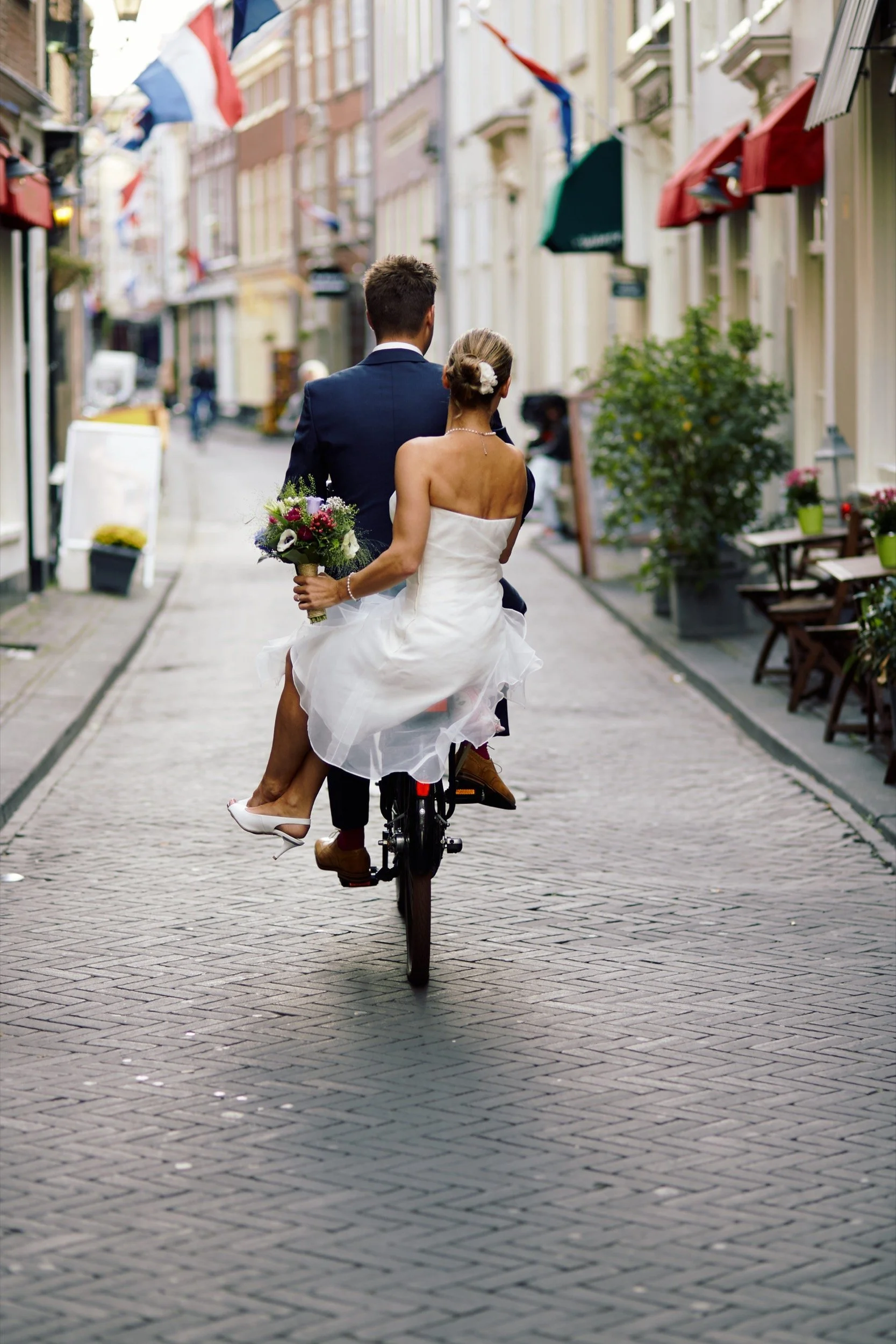 Een koppel op een fiets in een straat, waarbij de vrouw een bruidsjurk draagt met een bloemenboeket, en de man een pak, tijdens een huwelijksfeest.