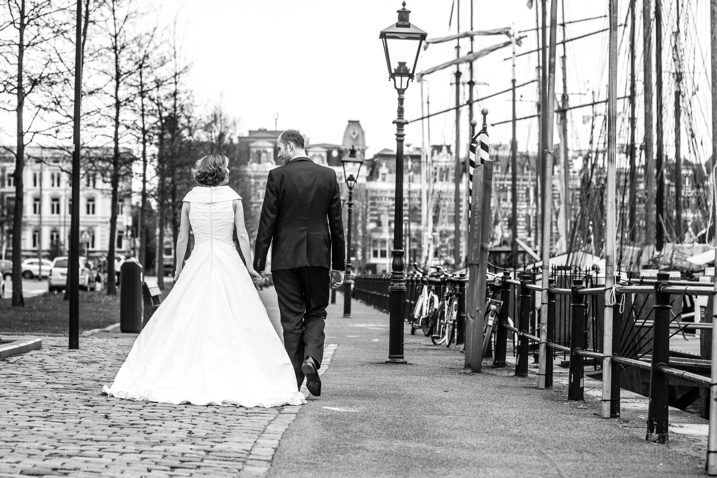 Een bruid en bruidegom lopen hand in hand langs een kade met boten en schepen, de foto is zwart-wit.