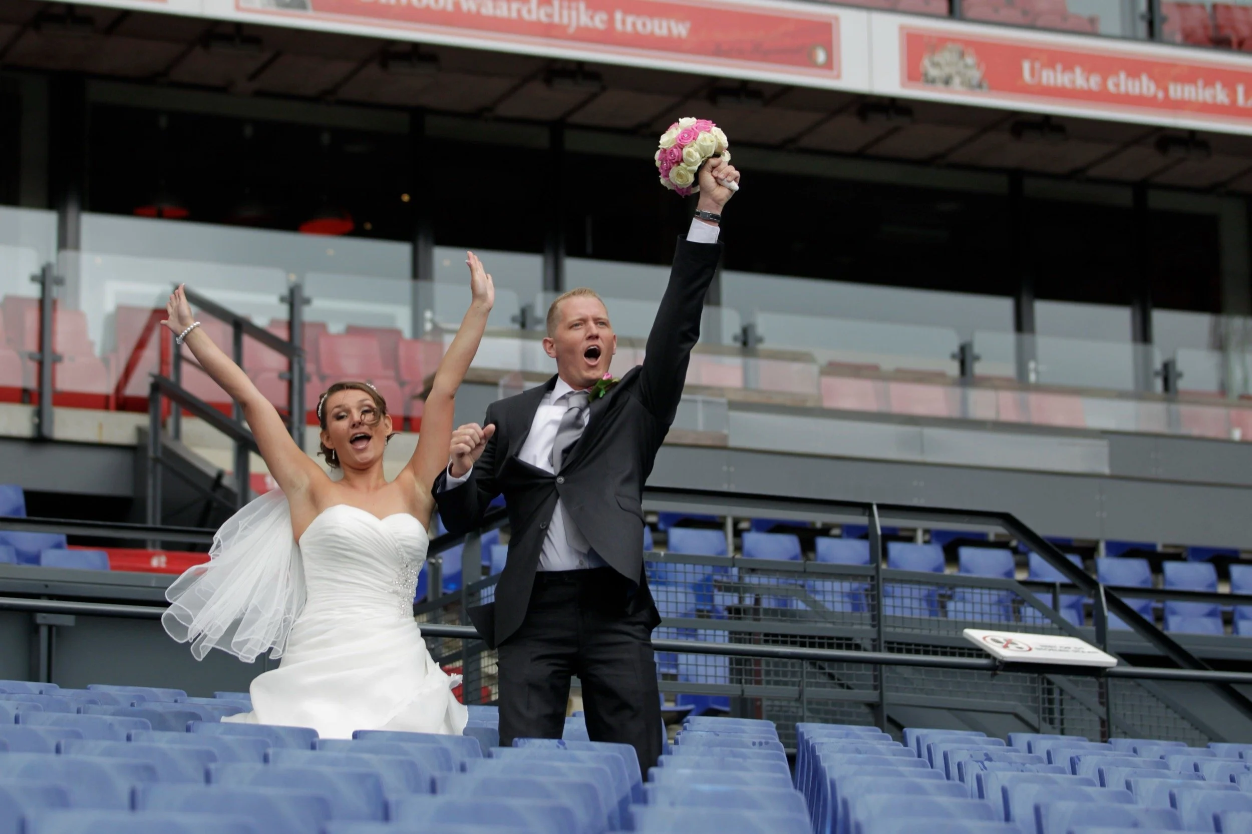 Een bruid en bruidegom vieren samen op een lege sporttribune, de bruidegom hef een boeket omhoog en beiden lijken blij en opgetogen.