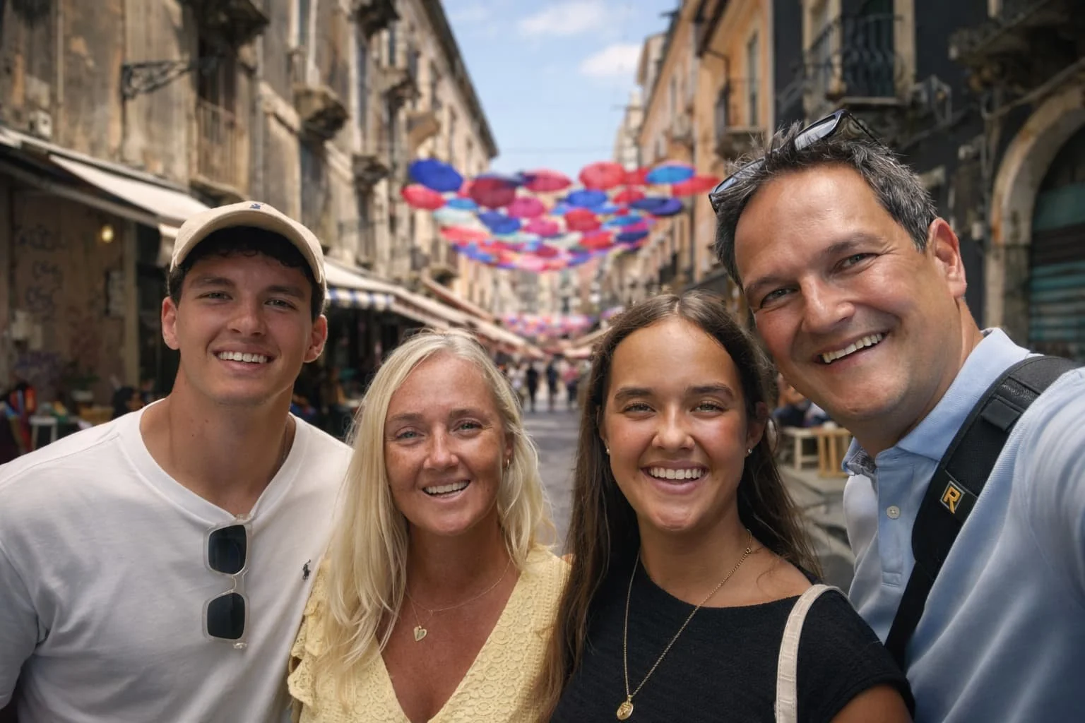 Vijf lachende mensen maken een selfie op een straat met kleurrijke parasols boven hen.