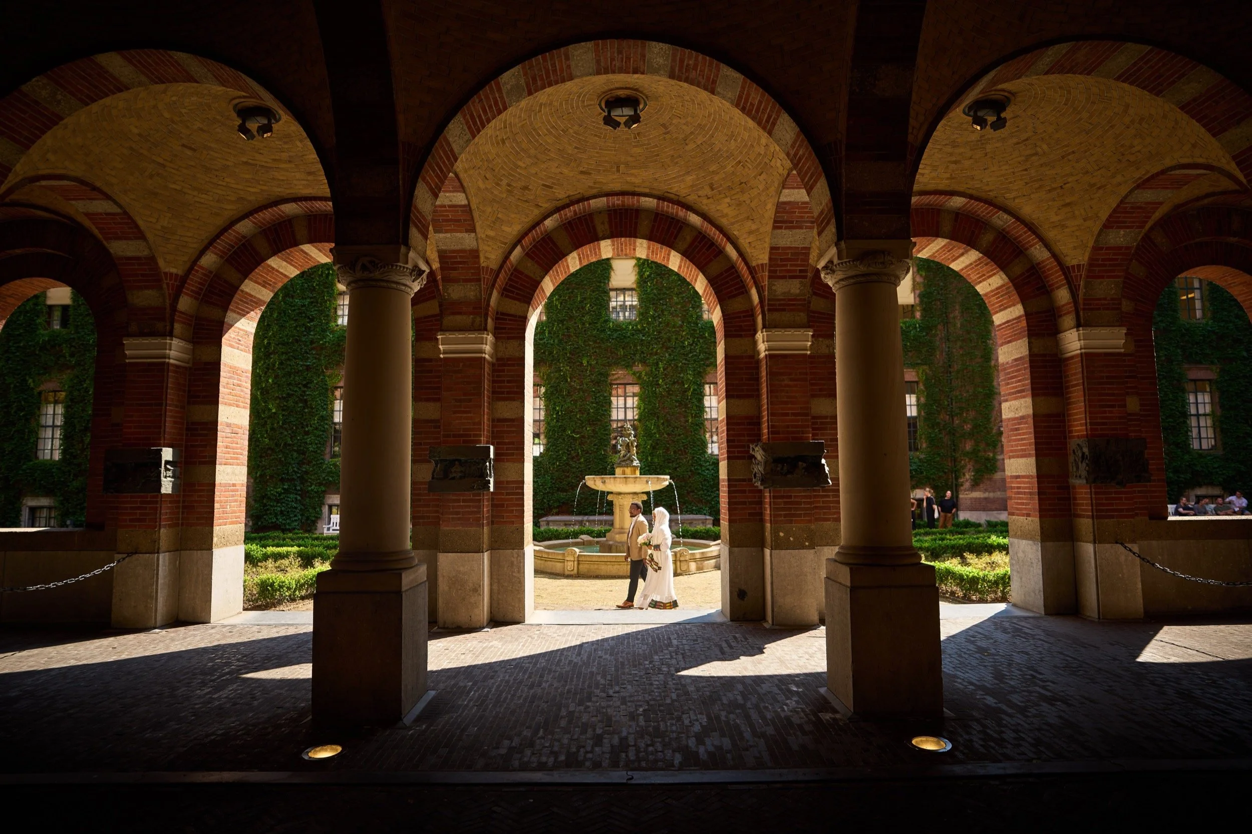 Twee mensen wandelen door een arcadewoning met een fontein in de tuin, omgeven door groene klimplanten op de achtergrond.