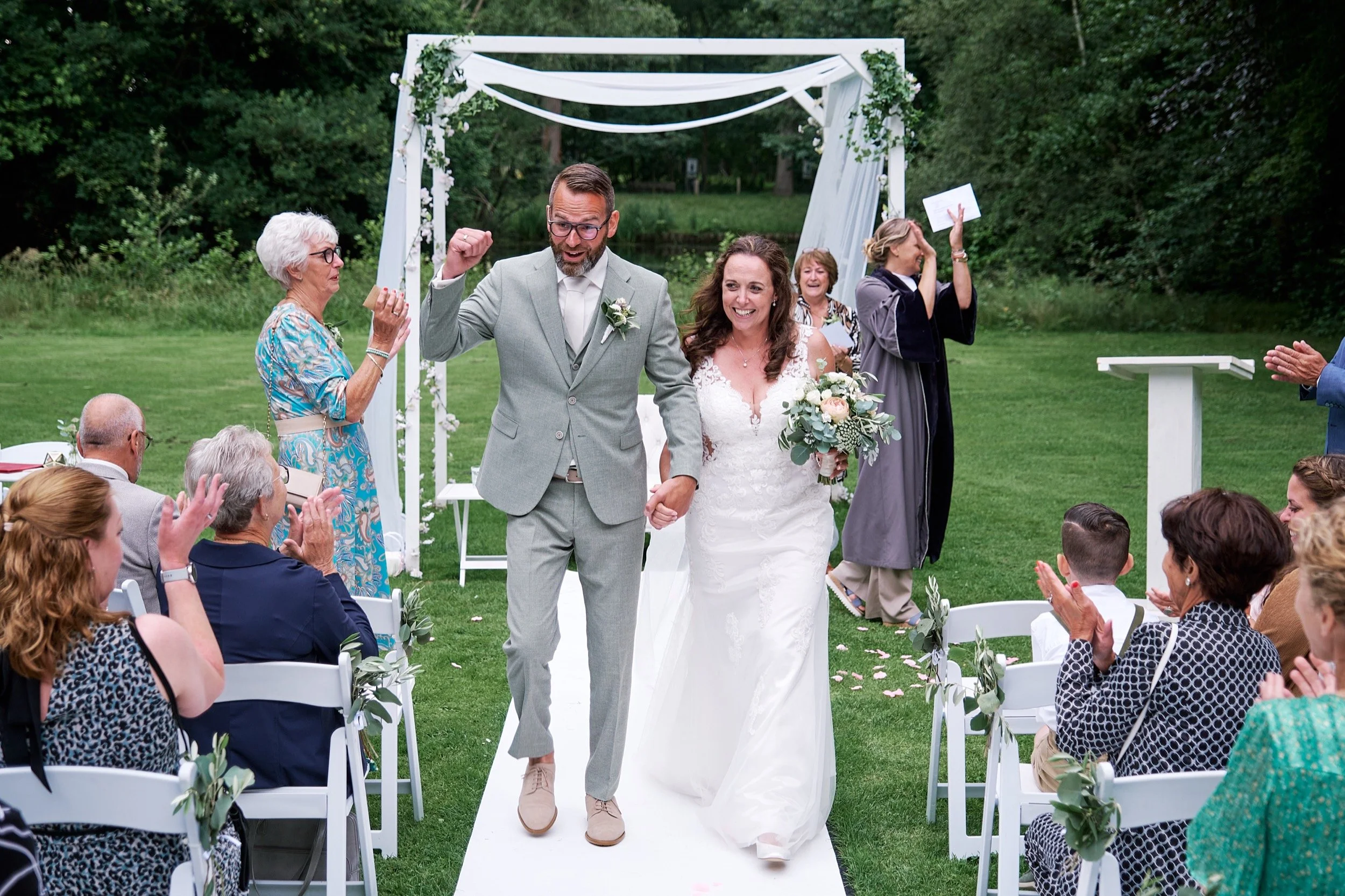 Gezicht op een huwelijksceremonie met een getrouwd koppel dat de huwelijksgeloften uitspreekt, omringd door gasten die applaudisseren. Er is een witte trouwgordijn en een groene tuin op de achtergrond.