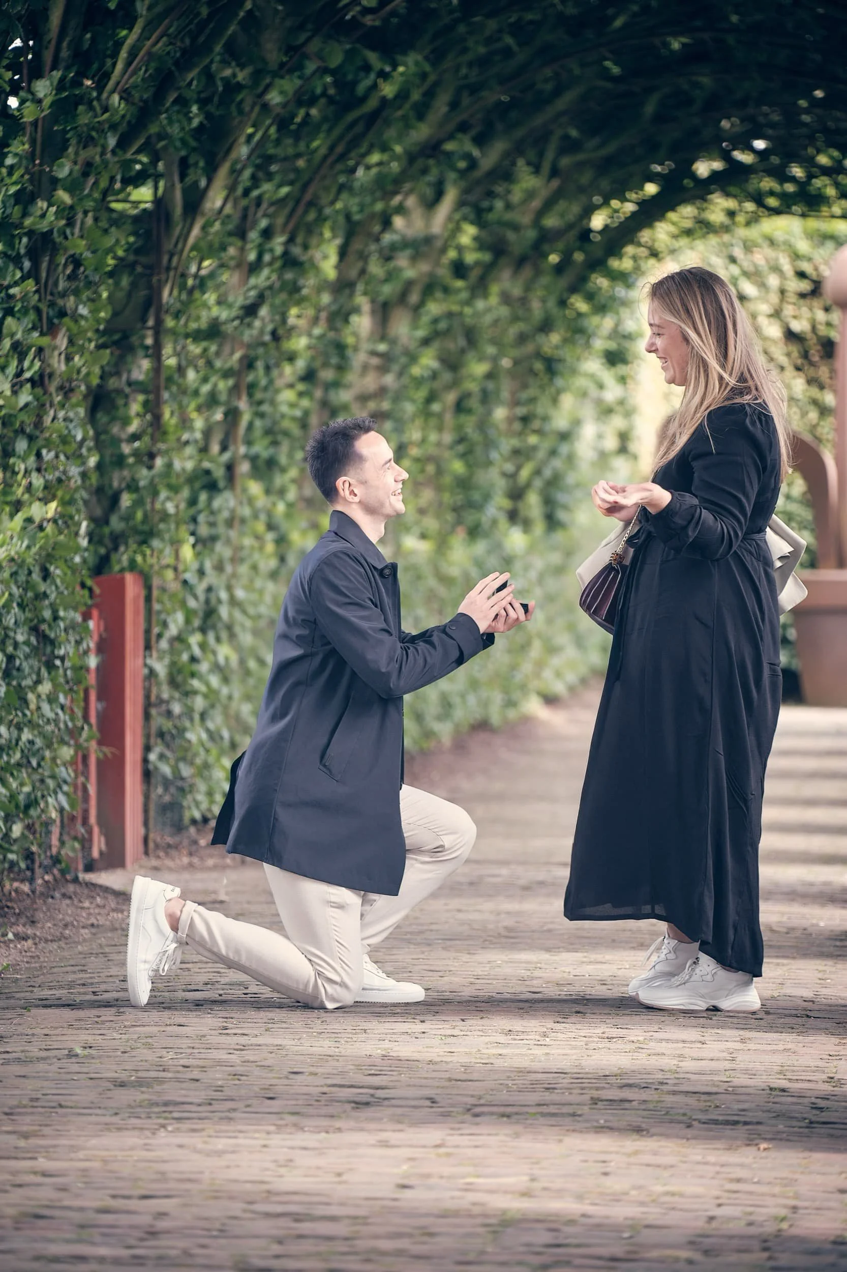 Man doet een voorstel terwijl hij op één knie ligt voor een vrouw in een park, beiden glimlachen.