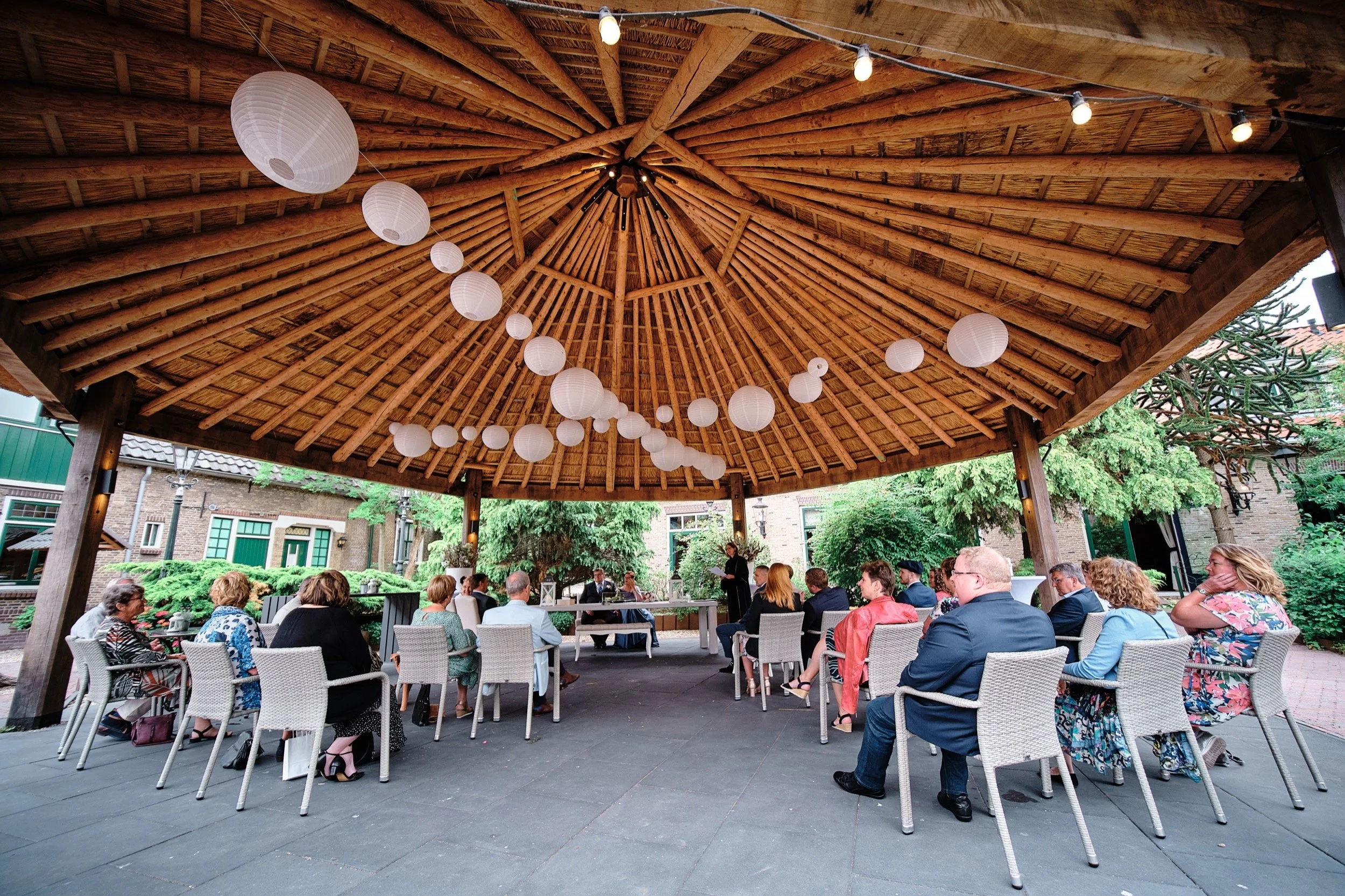 Een groep mensen zit onder een grote ronde houten pergola met een rieten dak, festoonverlichting en witte papieren lampionnen. Ze nemen deel aan een bijeenkomst of ceremonie buiten, omringd door groene bomen en een gebouw met grote ramen.
