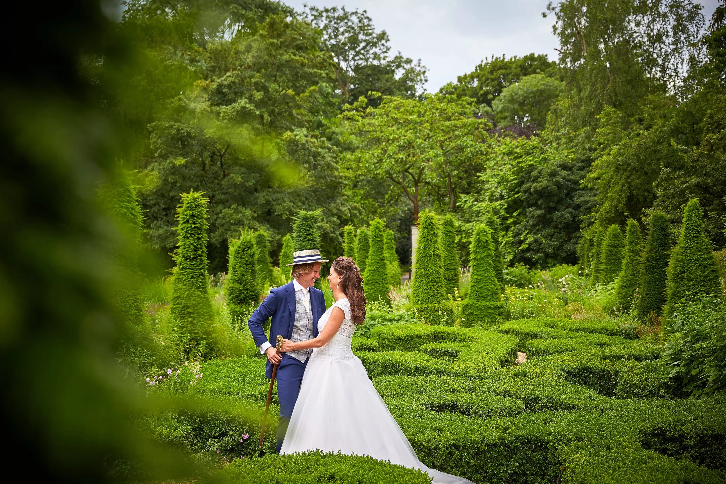 Een bruid en bruidegom in een groene tuin, dicht bij elkaar, lachen en kijken elkaar lief aan.