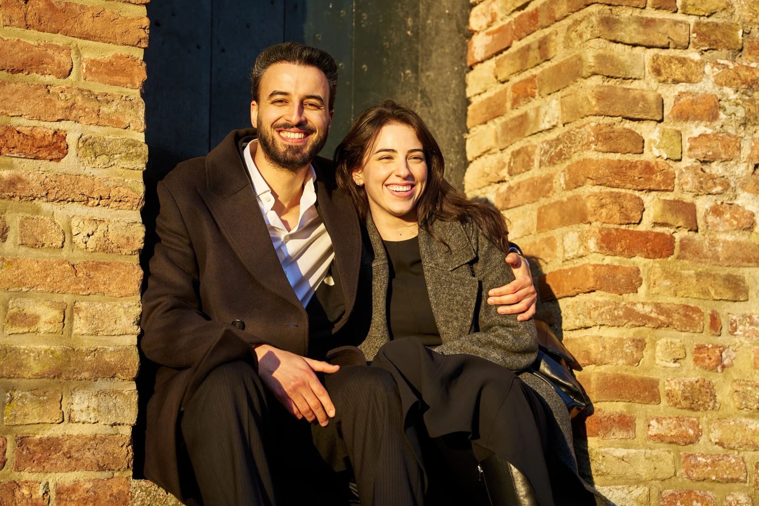 Twee lachende mensen zitten samen tegen een bakstenen muur in de zon, een man met een donkere jas en een vrouw met een grijze jas, omarmend en plezierig.