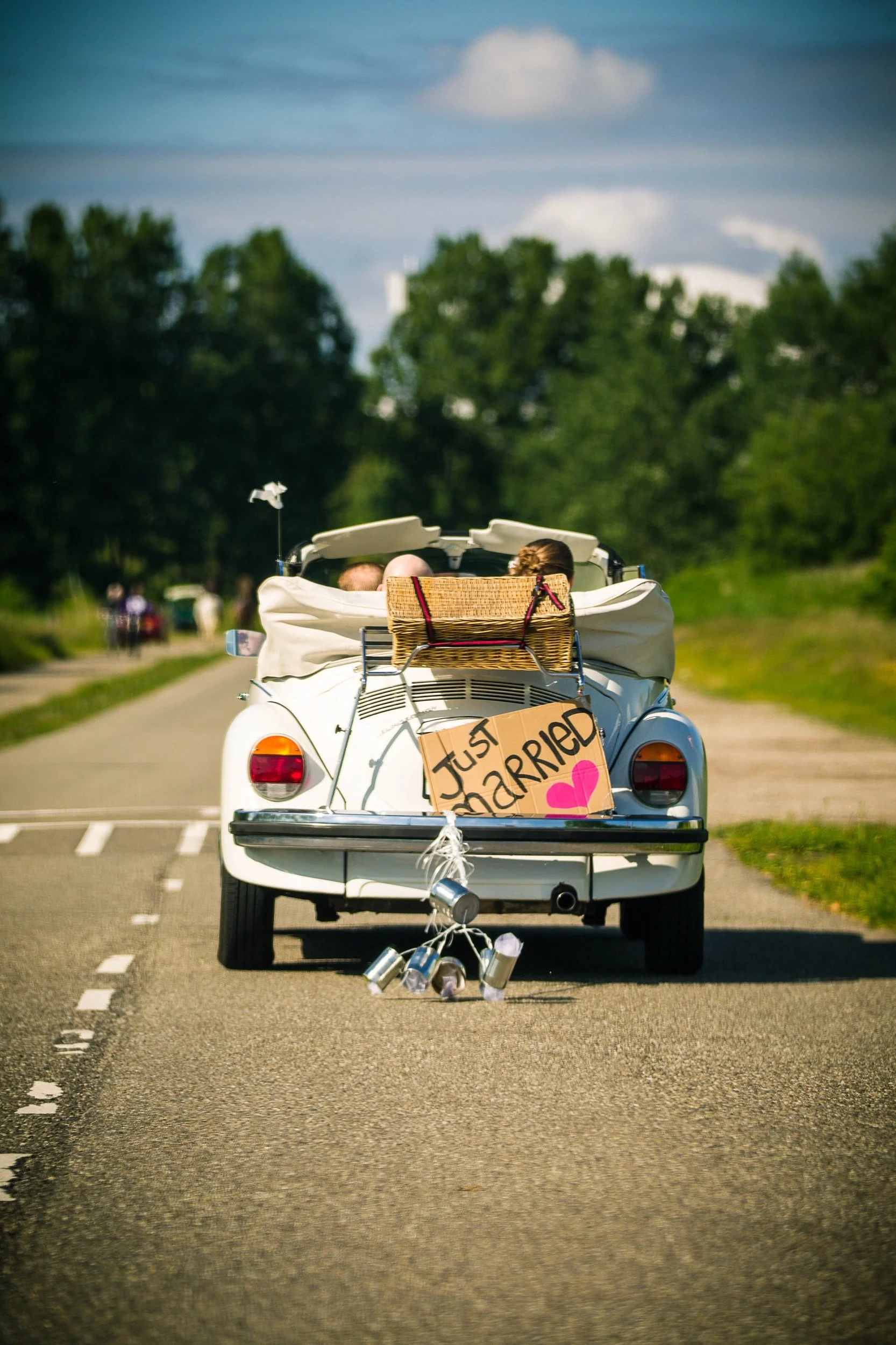 Een witte auto rijdt op een landweg met een bushokje rechts. Op de achterkant van de auto hangt een bord met de tekst 'Just married' en een roze hartje, met aan de bumper draden met ingeschroefde blikken.