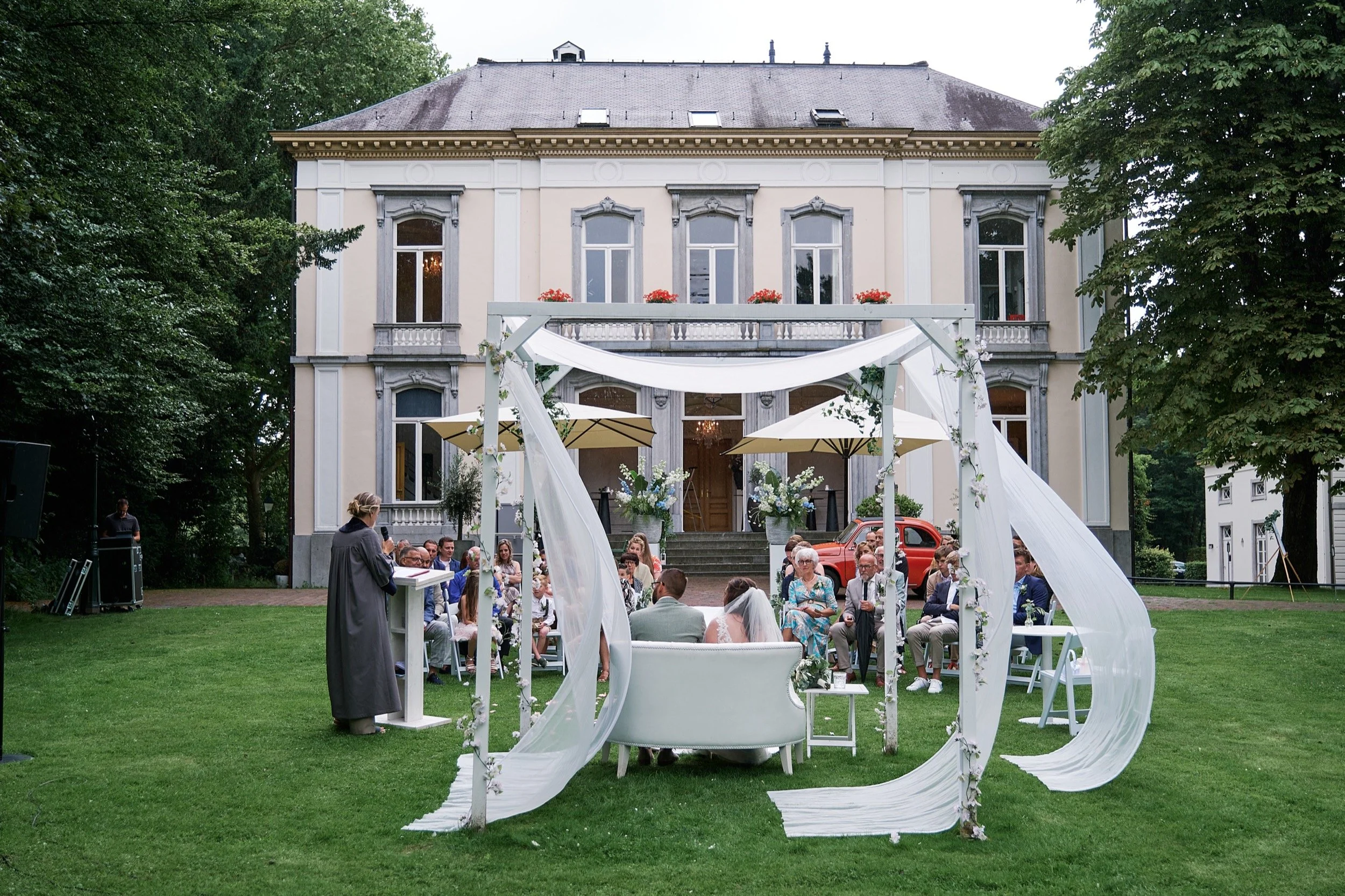 Een buitenhuwelijk met gasten zittend in de tuin van een groot, wit balkonhuis, een ceremoniële setting met een witte stoel, een witte bloemenbank en witte draperieën, en een ceremoniemeester die voor de gasten spreekt.