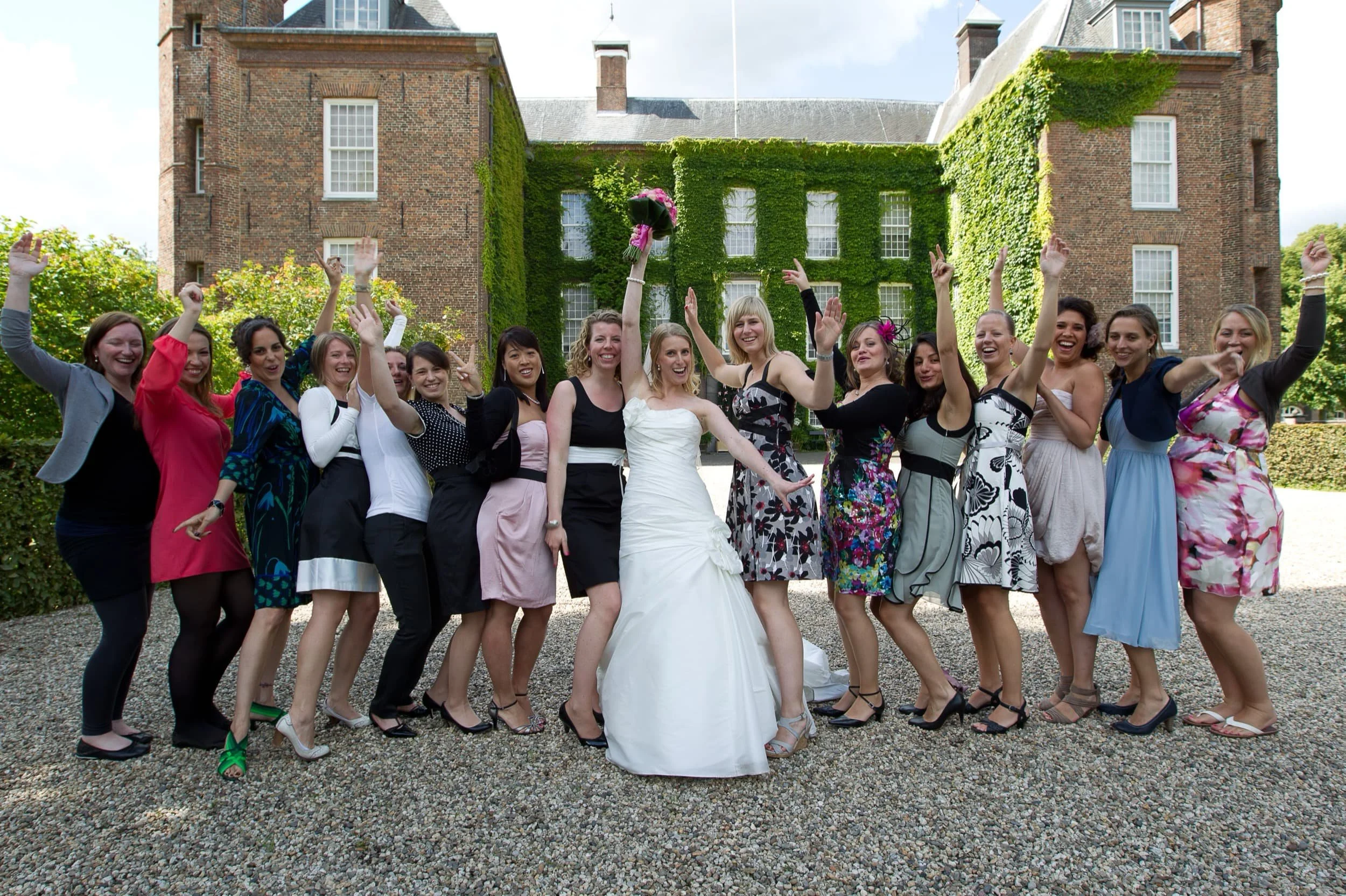 Een groep vrouwen in feestkleding die juicht en poseert voor een oude kasteel-achtige gebouw met groene klimop en ramen.