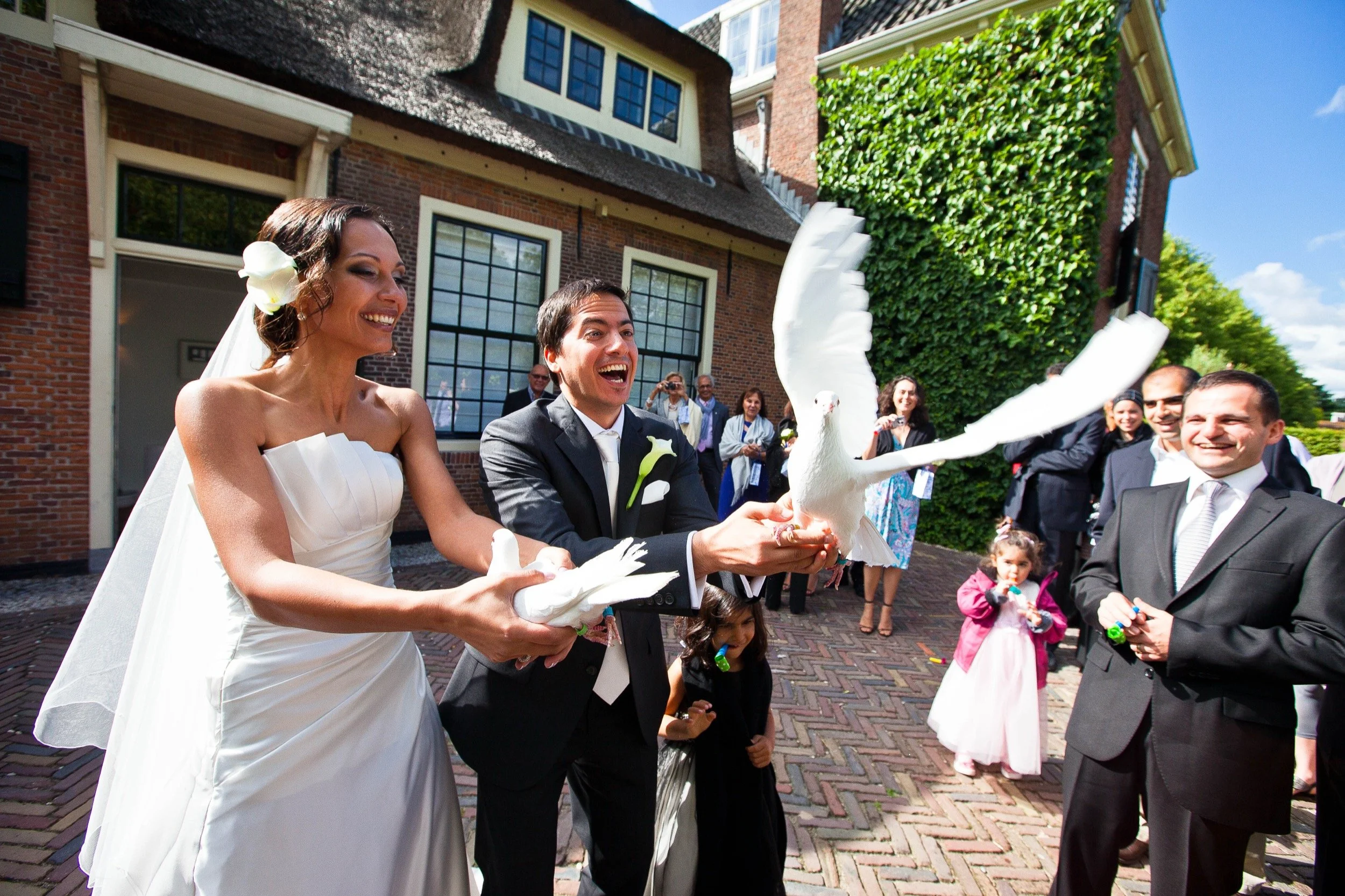 Een bruid en bruidegom houden samen een witte duif vast, die vleugels uitslaat, terwijl ze op hun huwelijksdag omringd door gasten lachen en genieten.