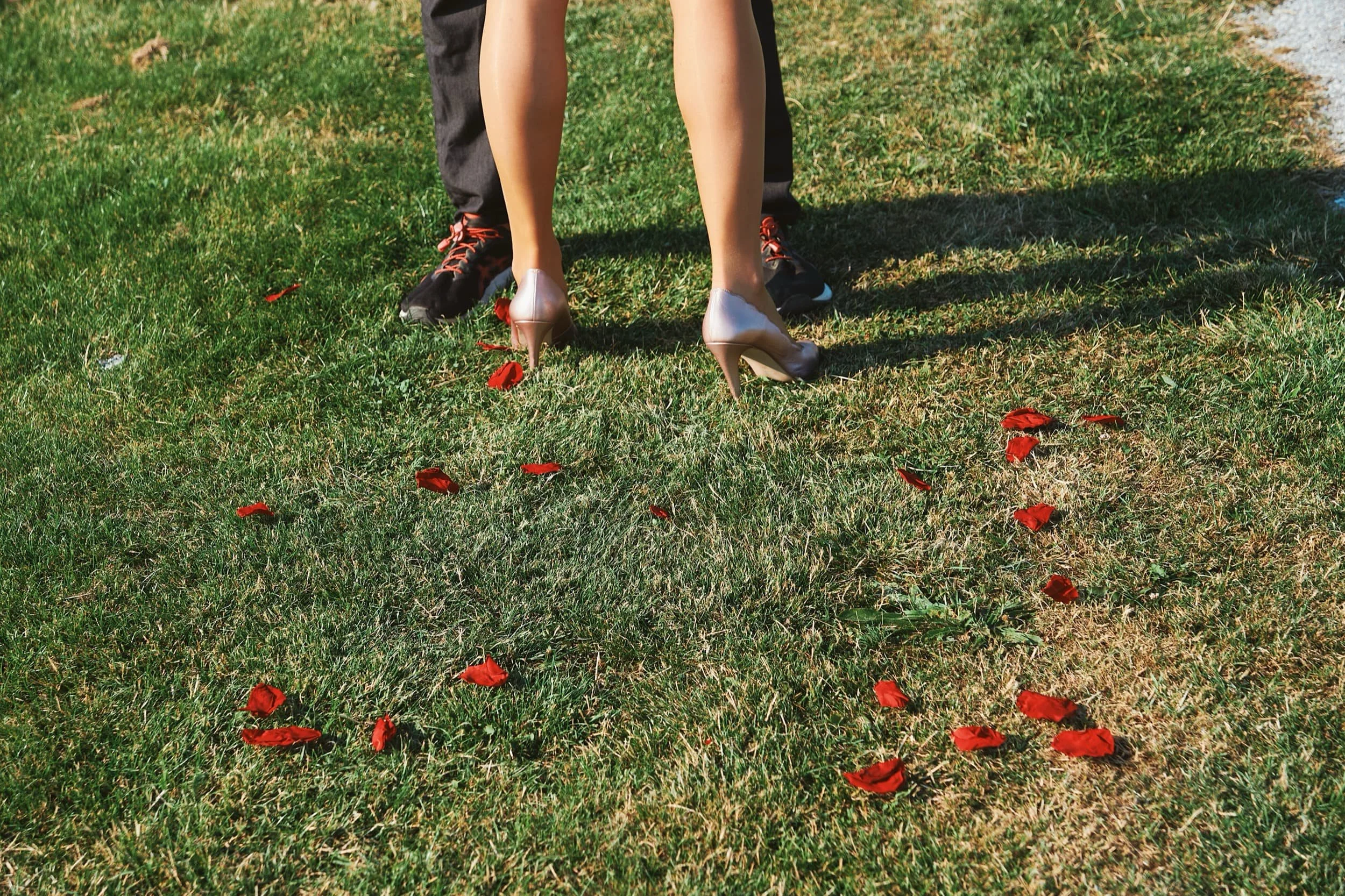 Twee mensen, een man en een vrouw, staan op gras met rode bloemenblaadjes. De vrouw draagt hoge hakken en de man sportieve schoenen.