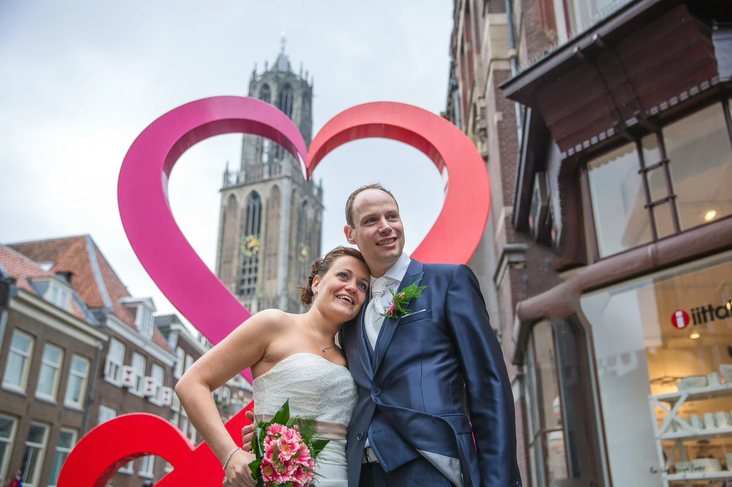 Een gelukkig bruidspaar poseert buiten met een grote roze en rode hartstructuur op de achtergrond, in een straat met historische gebouwen en een gotische kerk.