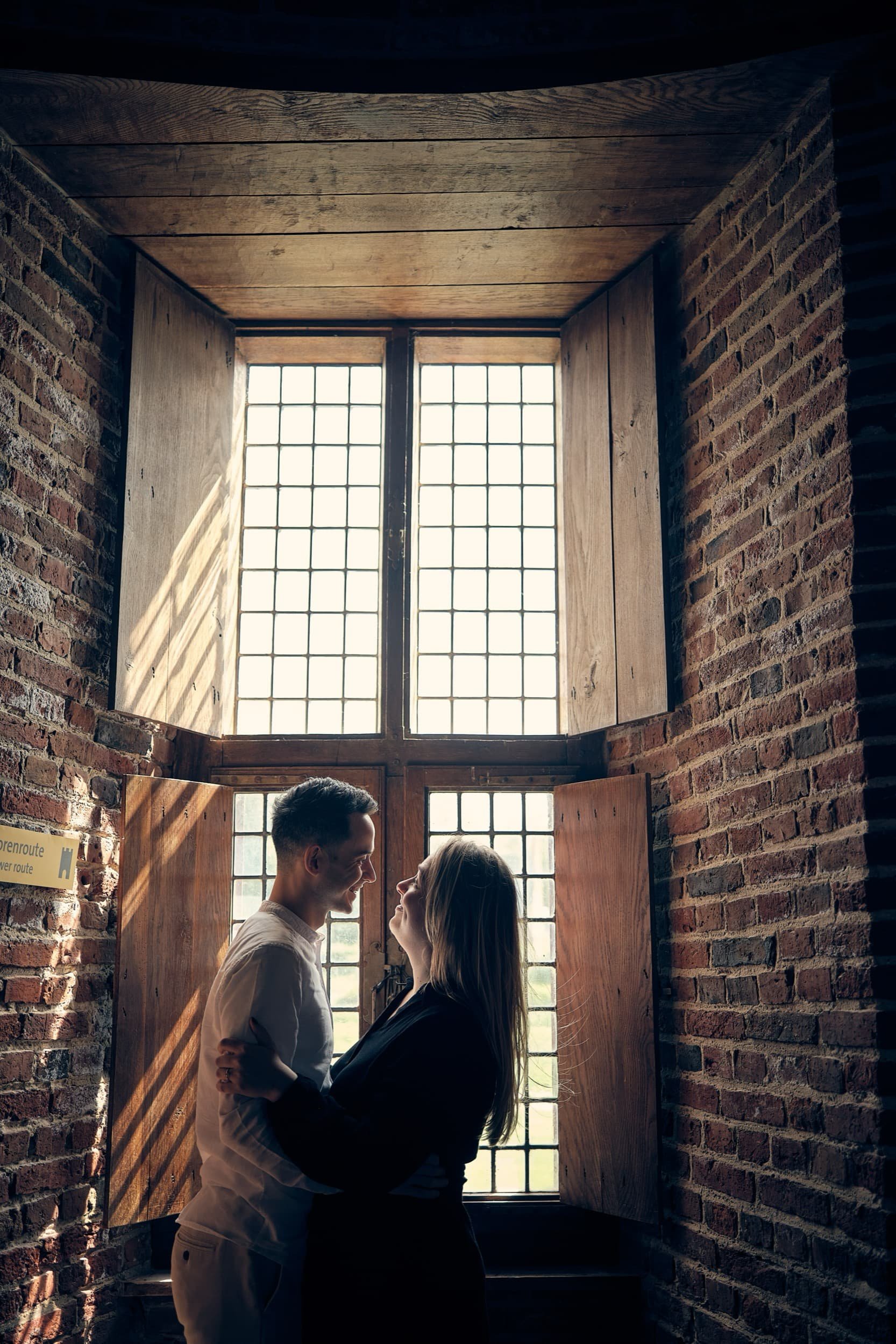 Twee mensen kijken elkaar liefkozend aan in een kamer met een groot, oude raampartij en bakstenen muren.