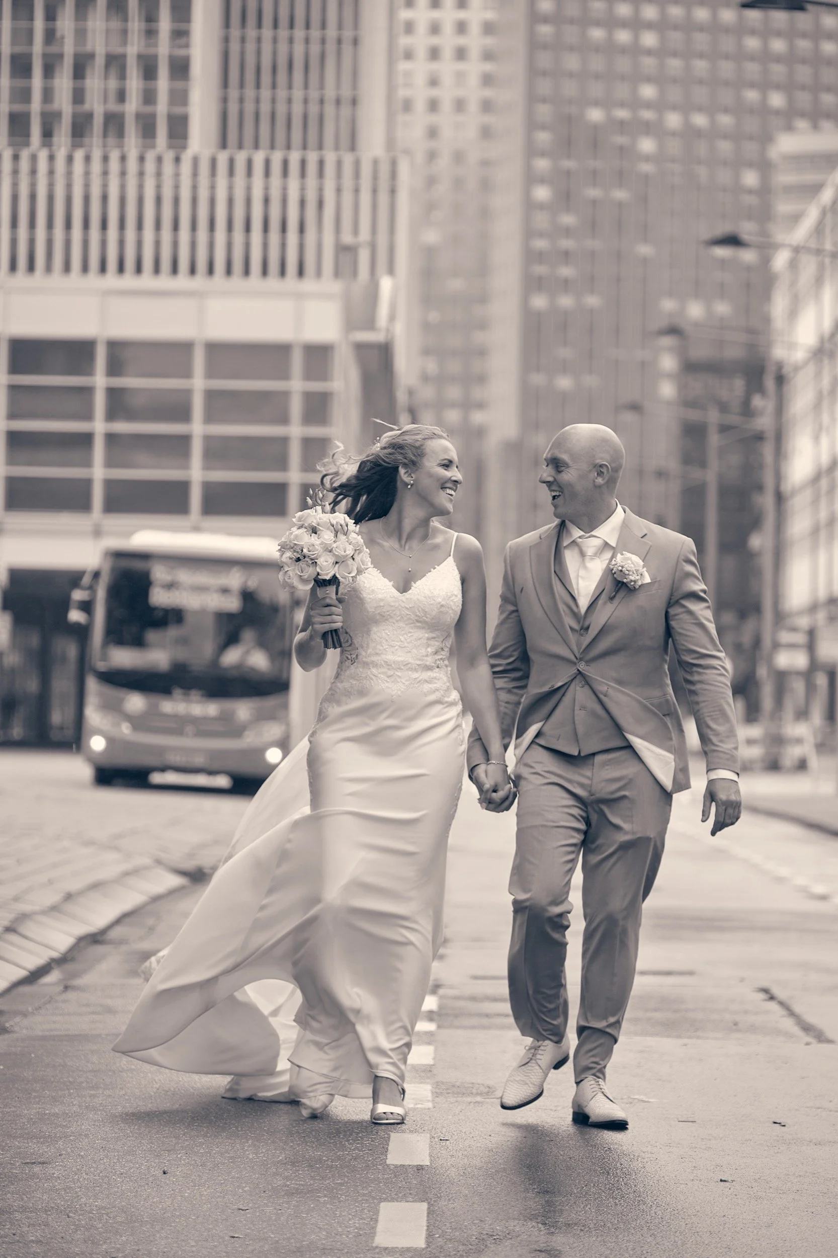 Een bruid en bruidegom lopen hand in hand door een stedelijk straatbeeld op hun trouwdag, beiden glimlachend.