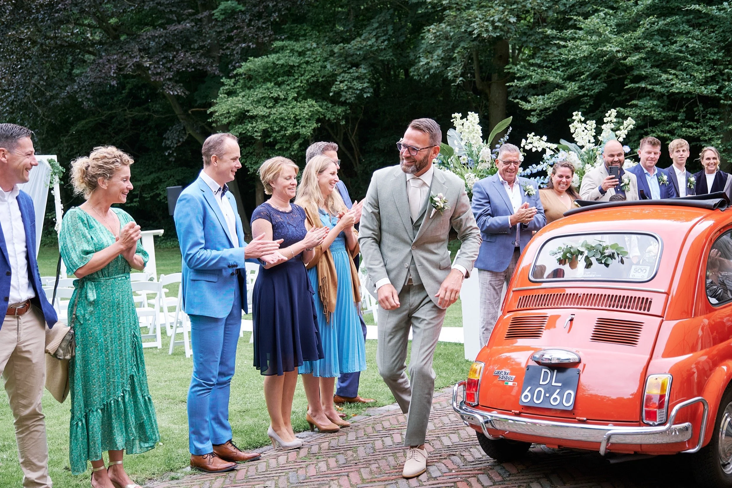Man in grijs pak loopt weg van een oranje vintage auto, omringd door lachende gasten tijdens een buitenceremonie, met groene bomen op de achtergrond.