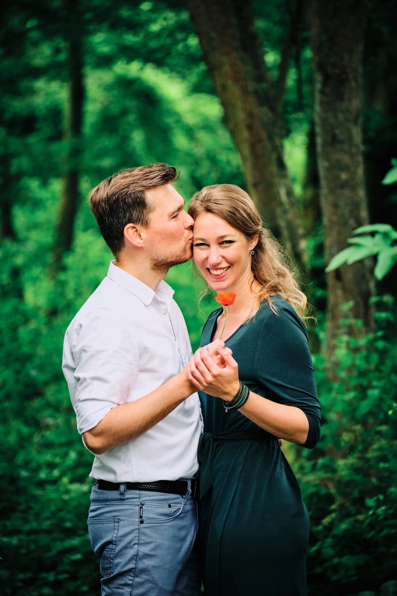 Een knuffelend paar in een bos, de man kust de vrouw op haar wang terwijl ze glimlacht, en ze houdt een oranje bloem in haar hand.