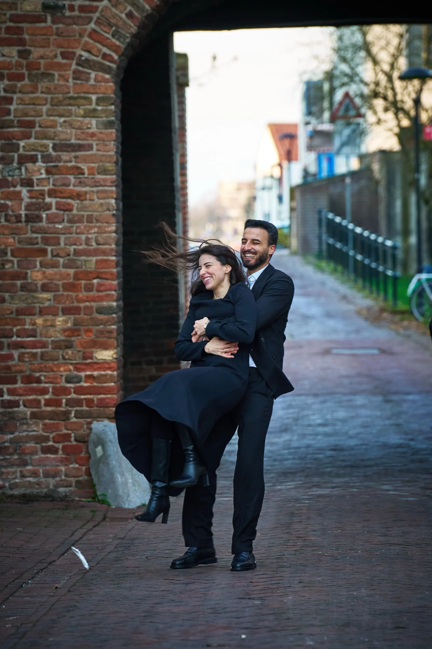 Een man tilt een lachende vrouw op in een straat met een stenen muur en een boog in de achtergrond.