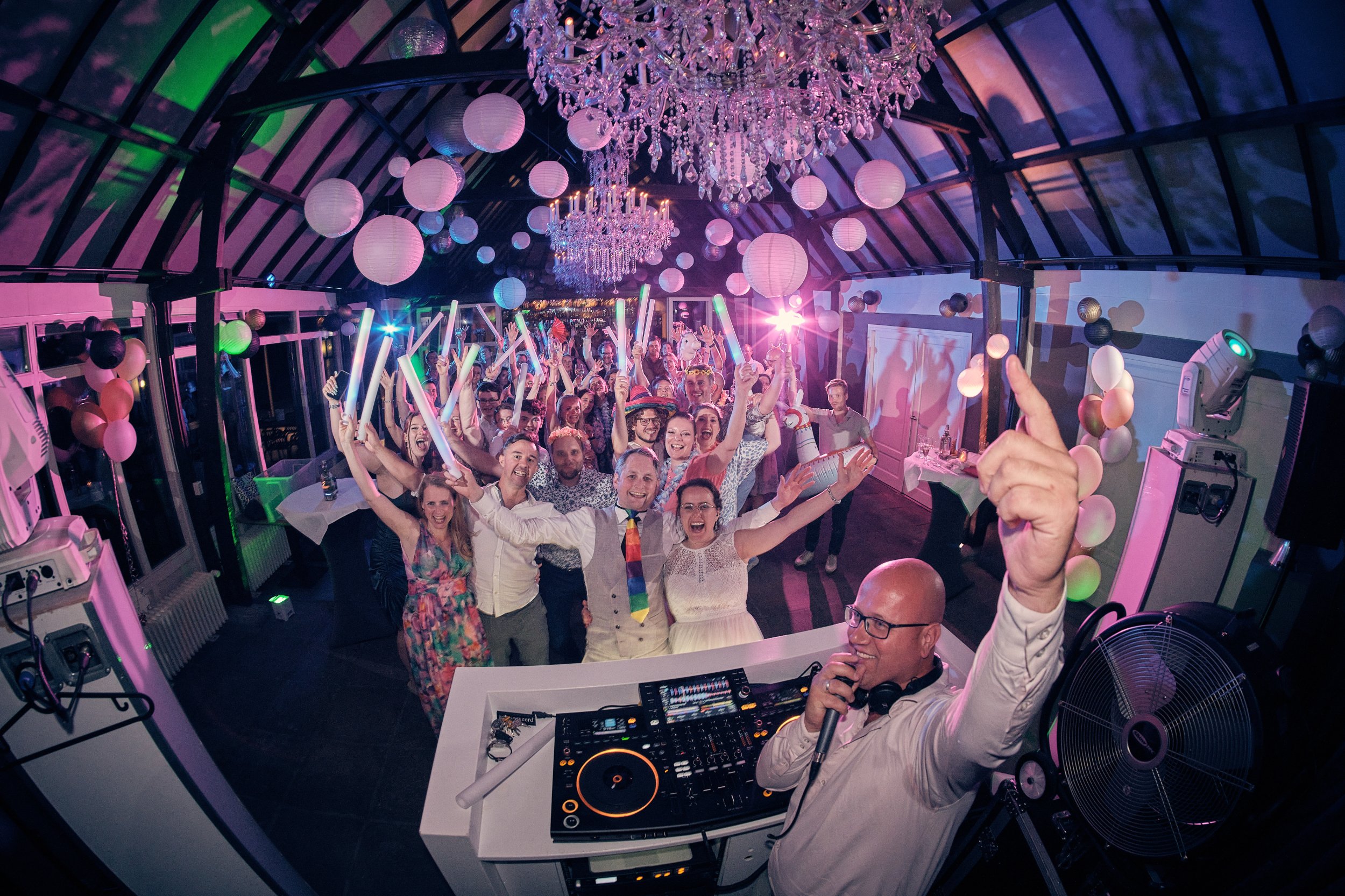 Bezoekers op een feest met kleurrijke lichtjes, ballonnen en chique verlichting in een serreachtig interieur, terwijl een dj op de voorgrond muziek draait.