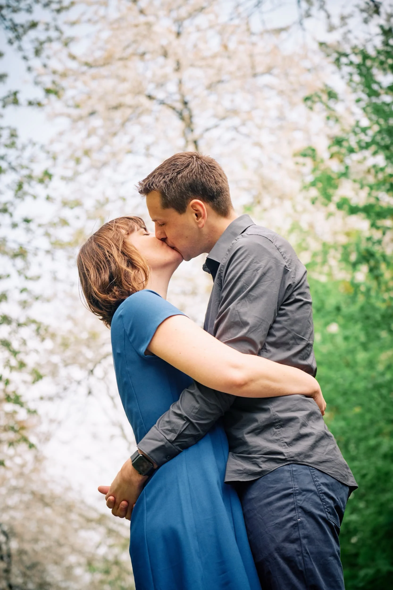 Een koppel deelt een kus in een park met bloesemende bomen.