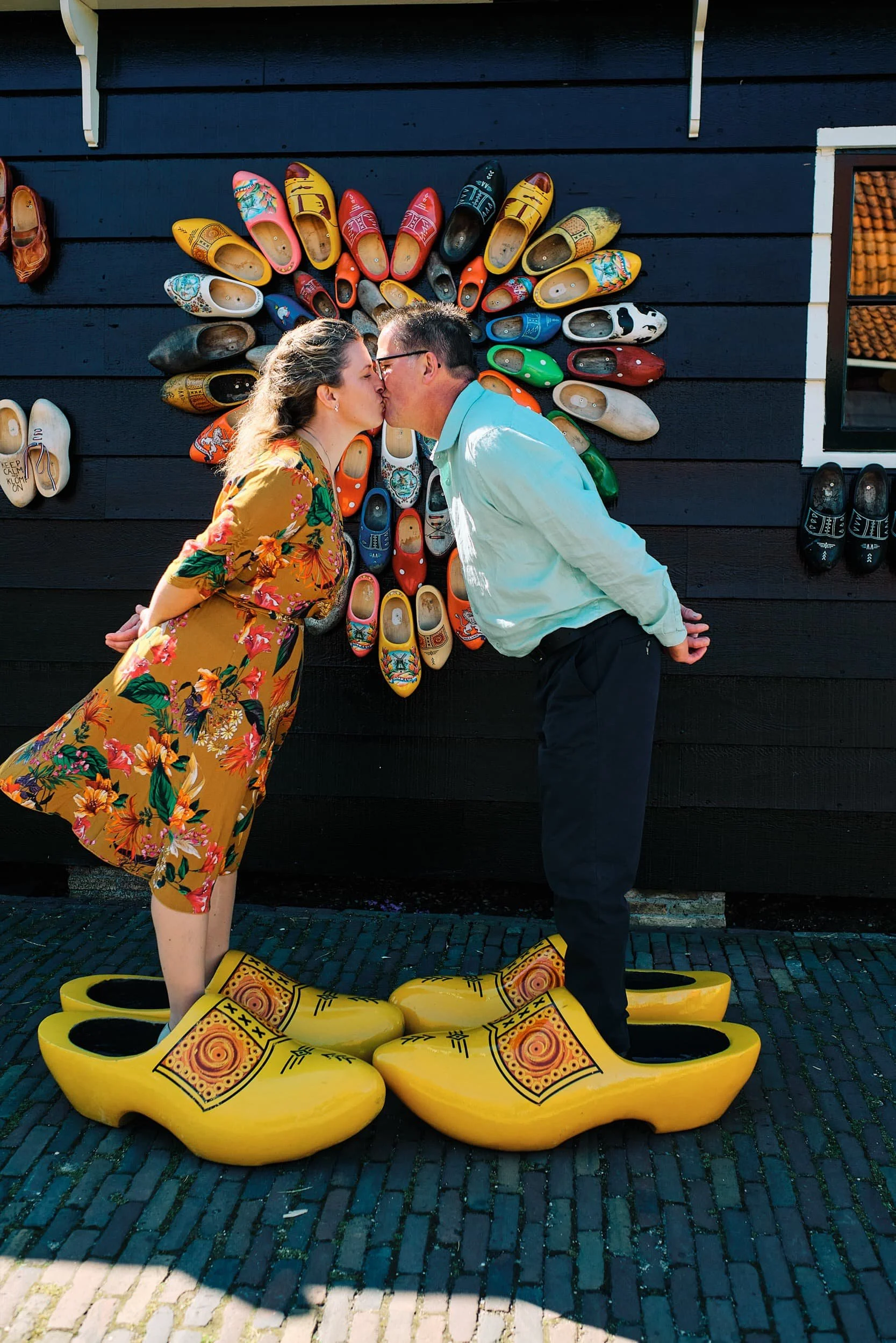 Een stel dat elkaar kust terwijl ze op grote gele klompen staan, voor een muur versierd met kleurrijke klompen.
