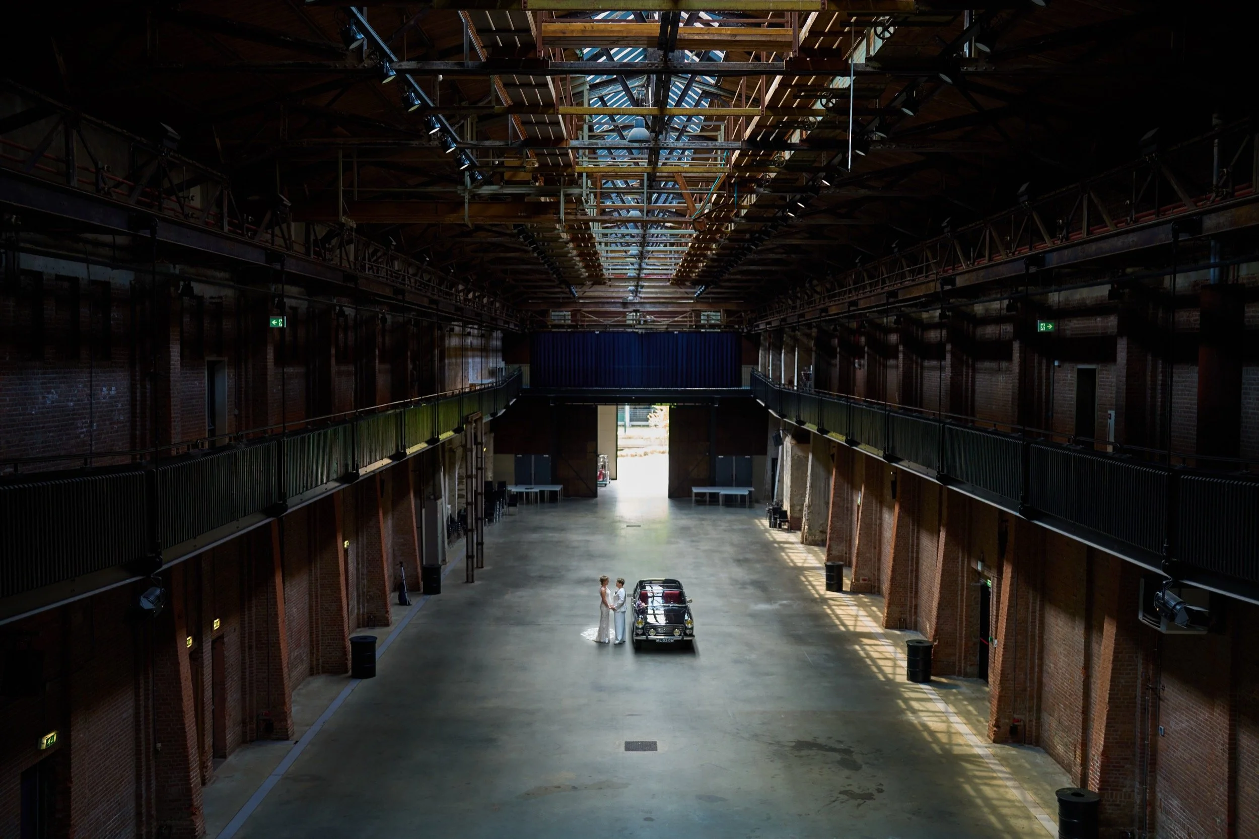 Een grote lege ruimte met hoge plafonds, twee vrouwen die in witte jurken hand in hand staan naast een zwarte auto.