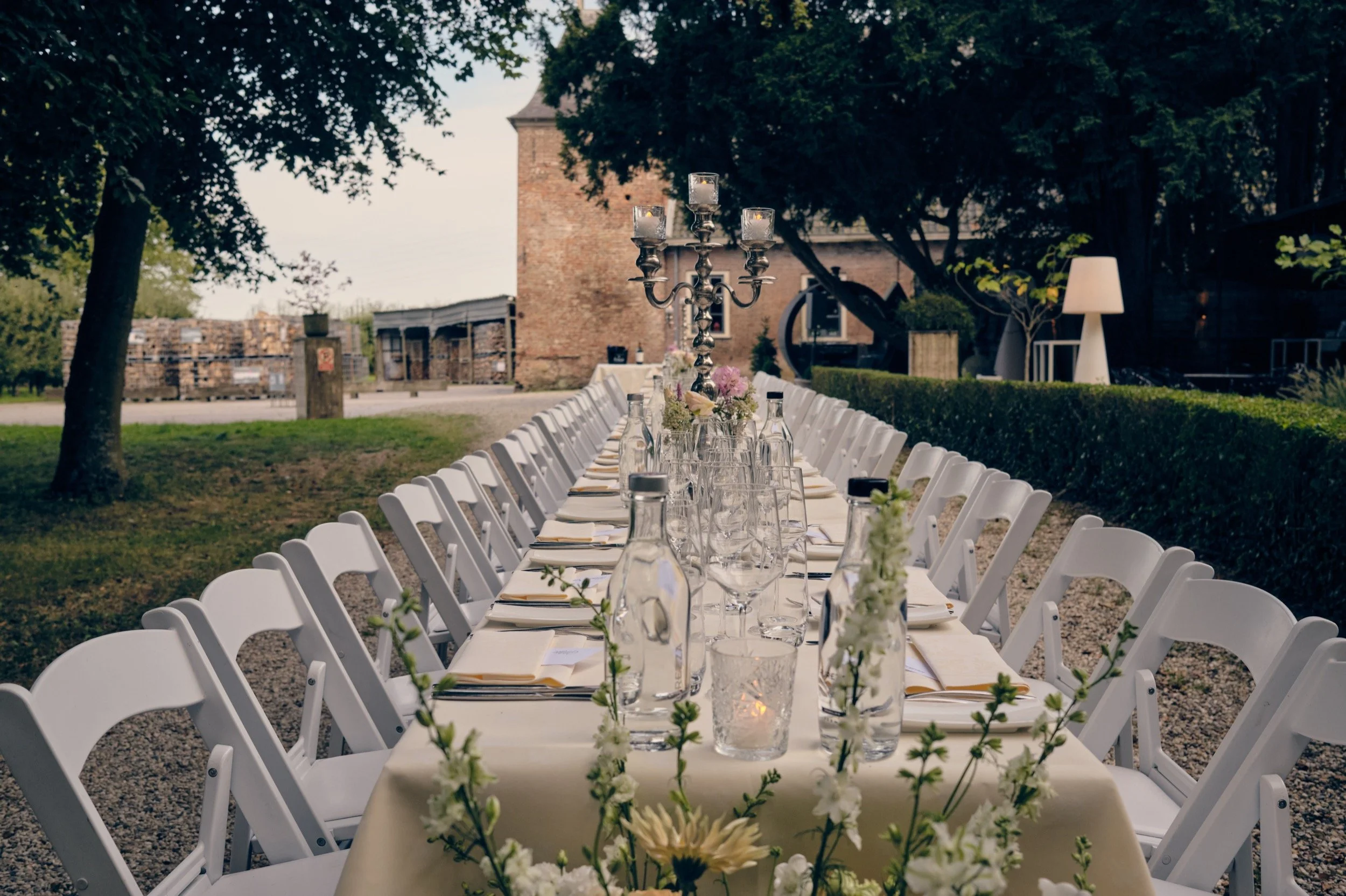 Lange feesttafel in een tuin met witte stoelen, een kroonluchter, bloemen en glazen, onder bomen en met een oude bakstenen toren op de achtergrond.
