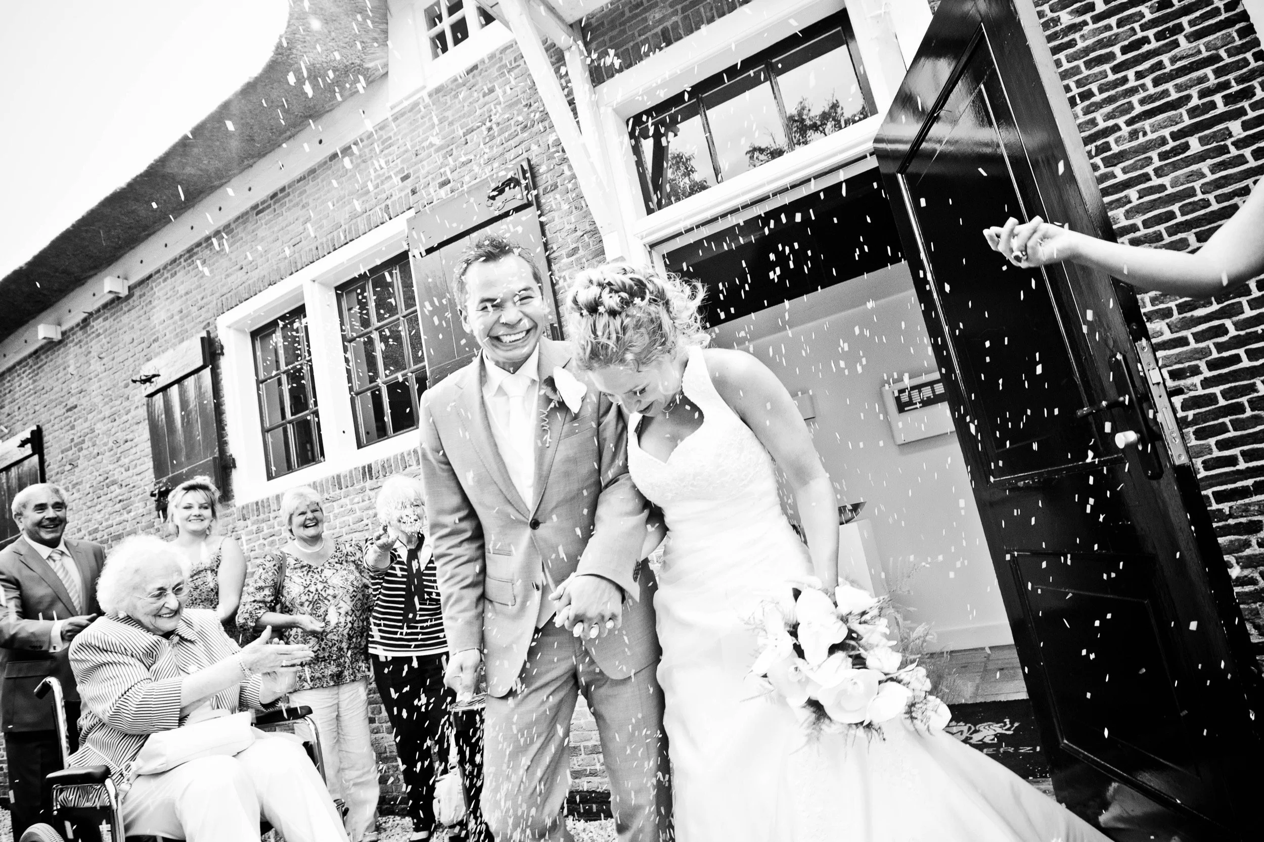 Bruid en bruidegom die samen door een confettiregen lopen, omringd door lachende gasten bij een bruiloft, buiten voor een bakstenen gebouw.