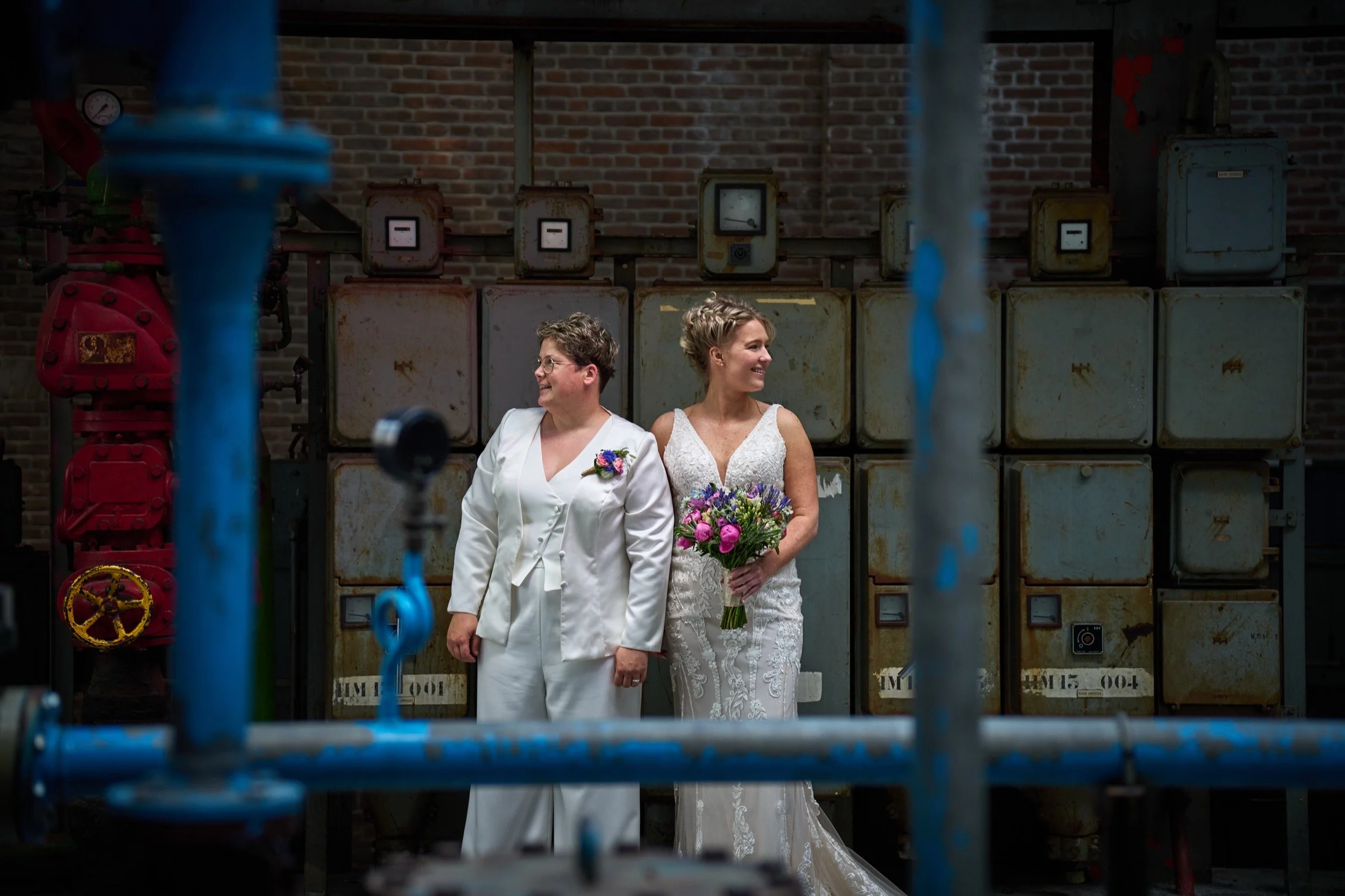 Twee vrouwen in witte bruiloftskleding staan zij-aan-zij voor een oude muur met meters, omringd door blauwe en rode industriële pijpen.