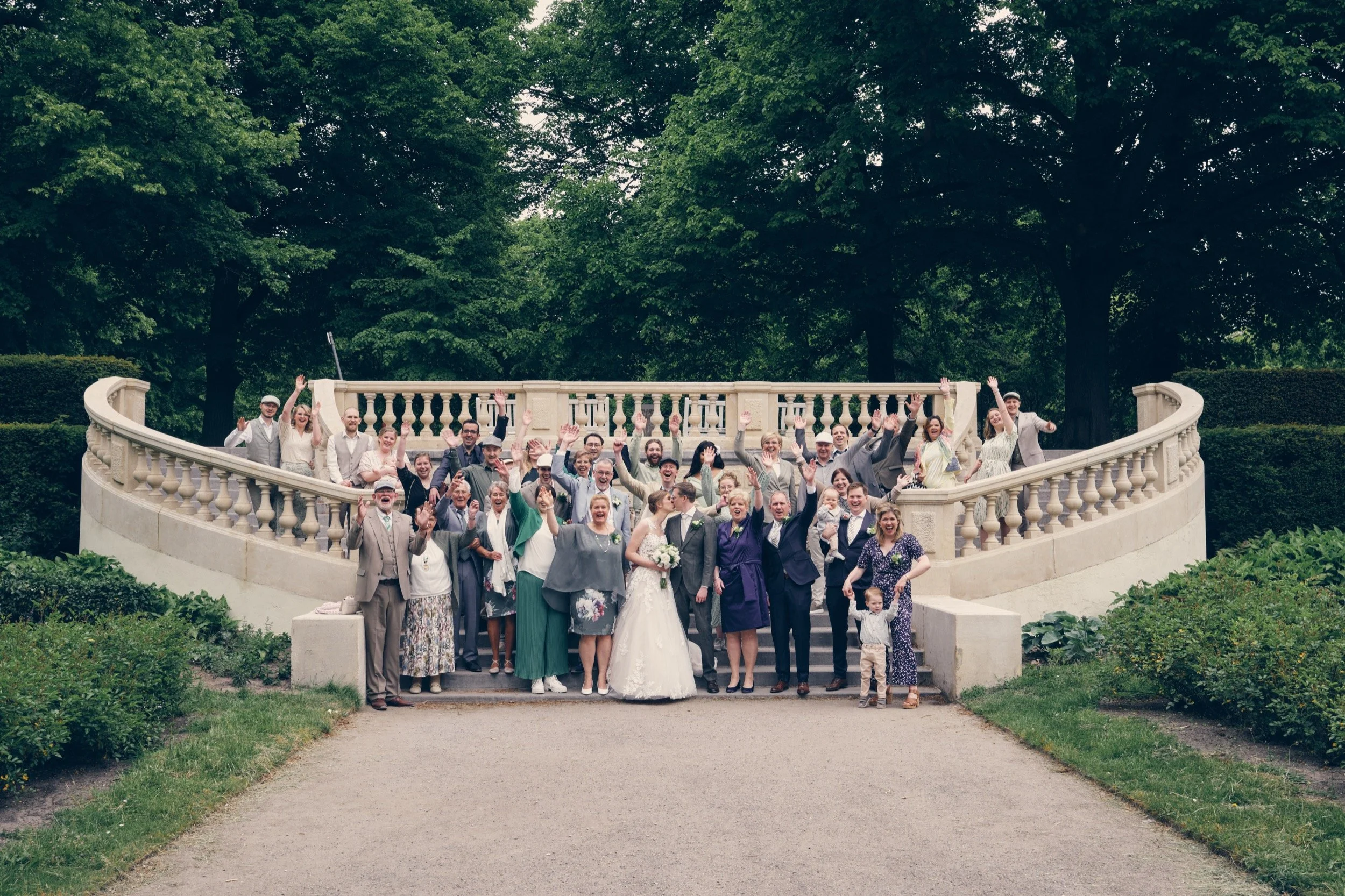 Een grote groep mensen die poseren voor een foto op een trappen in een park, waarschijnlijk tijdens een bruiloft.