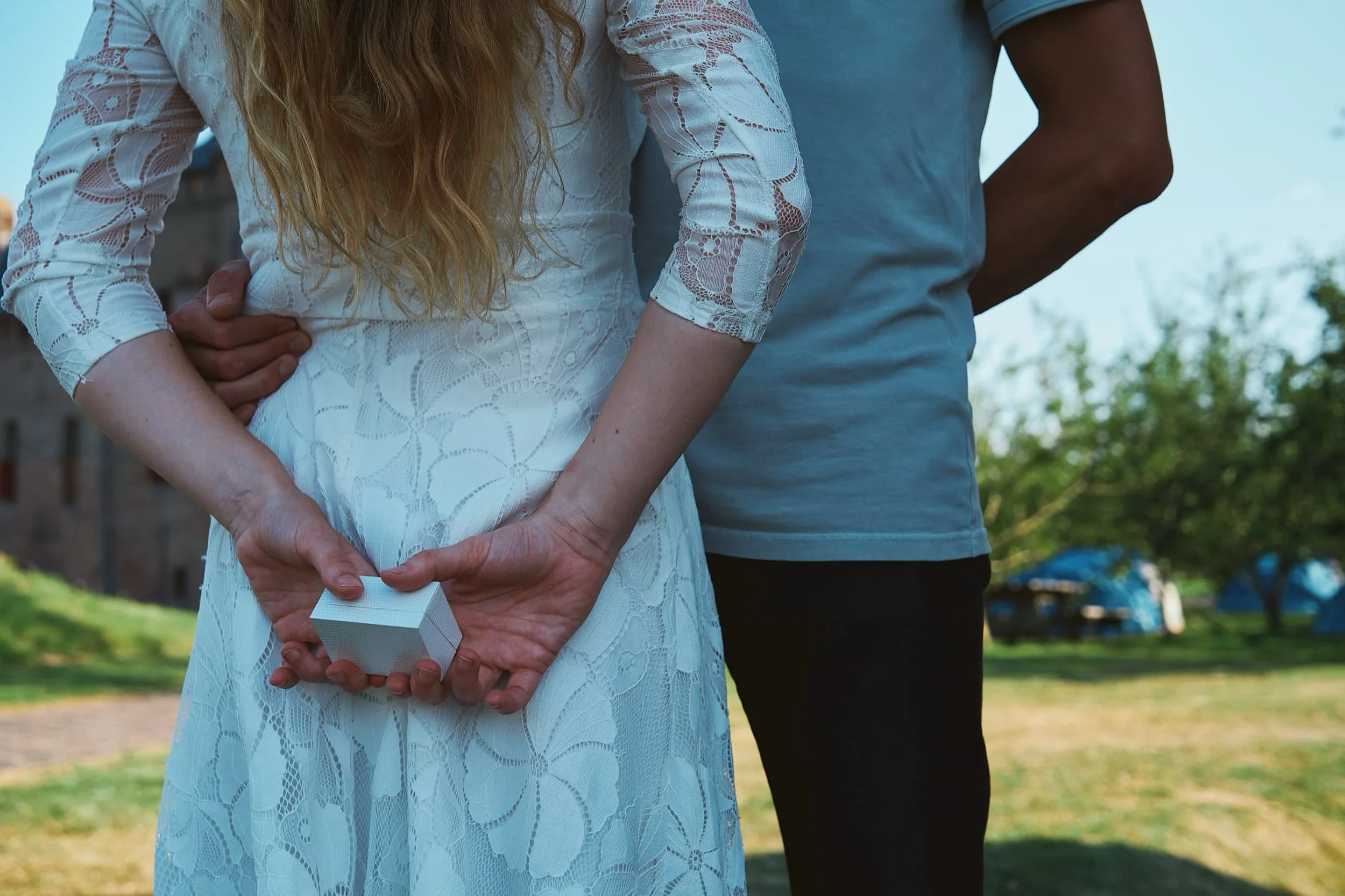 Een vrouw in een witte kanten jurk houdt een kleine witte doos achter haar rug, terwijl een man naast haar staat, beiden in een buitenomgeving.