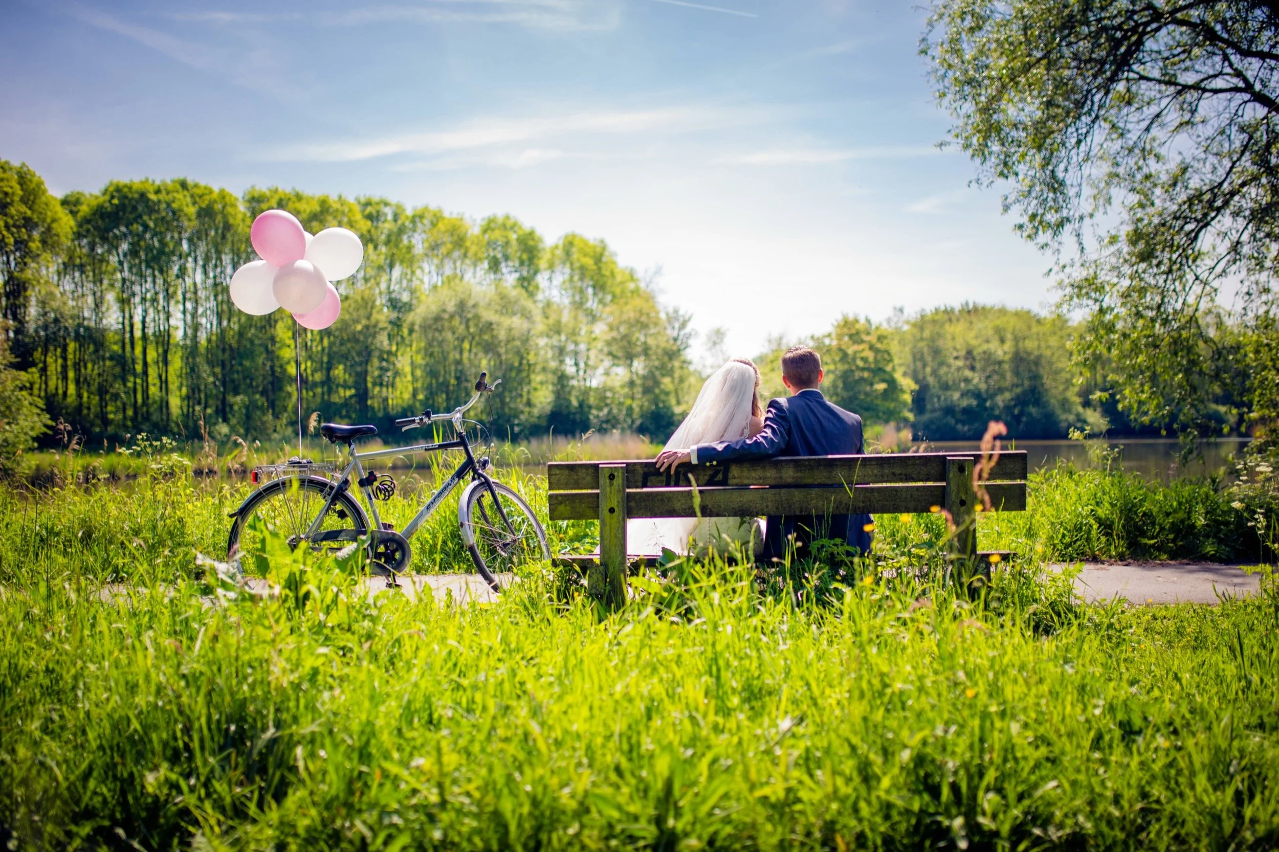 Een stelletje zit op een bankje naast een meer, omgeven door groen en bomen, met een fiets en ballonnen op het gras erbij.