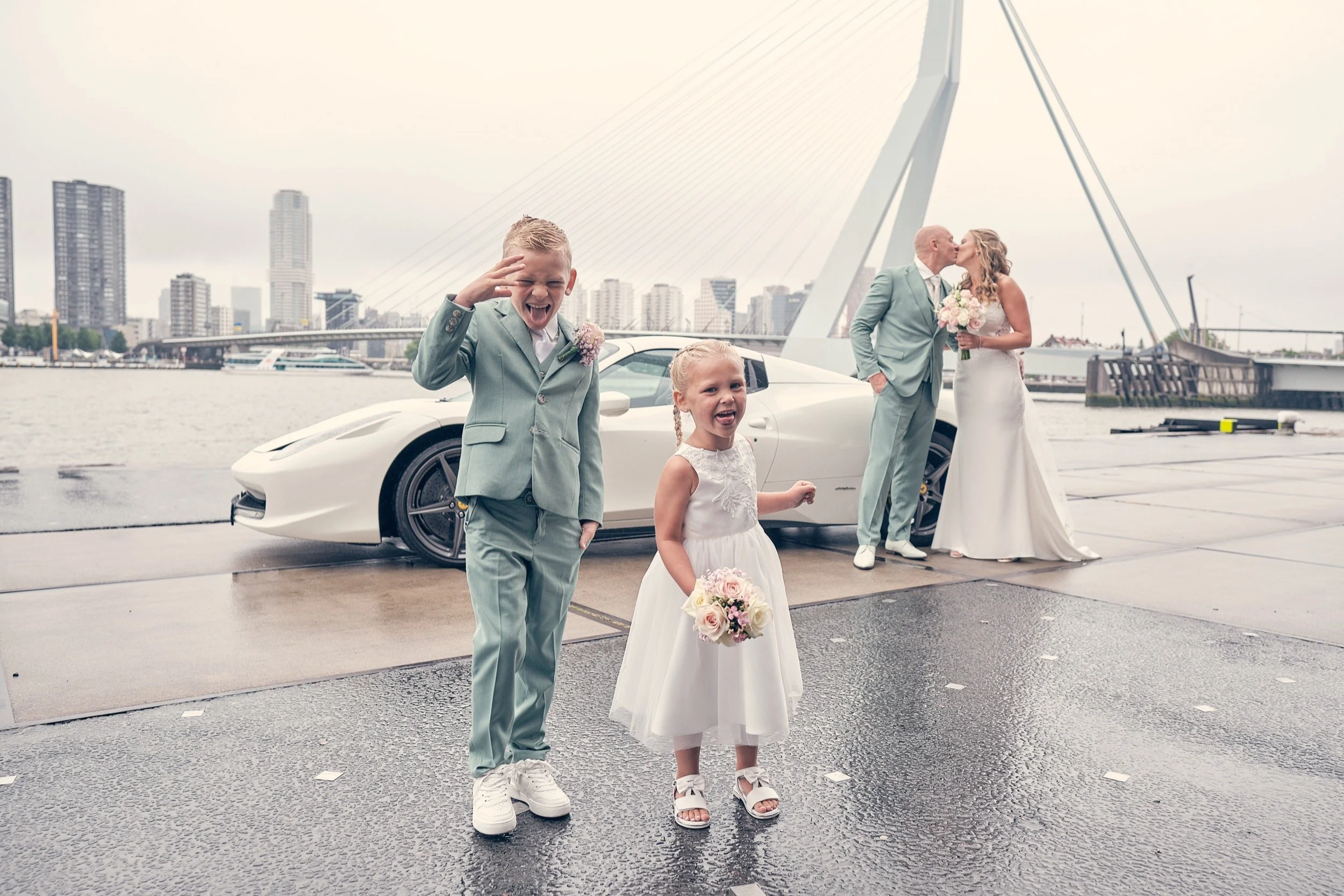 Een bruid en bruidegom kussen op een brug met een witte sportwagen op de achtergrond. Twee jonge kinderen, een jongen in een lichtblauw pak en een meisje in een witte jurk, staan vooraan, met de jongen die de camera uitdaagt en de meisje die in een jurk een boeket vasthoudt.