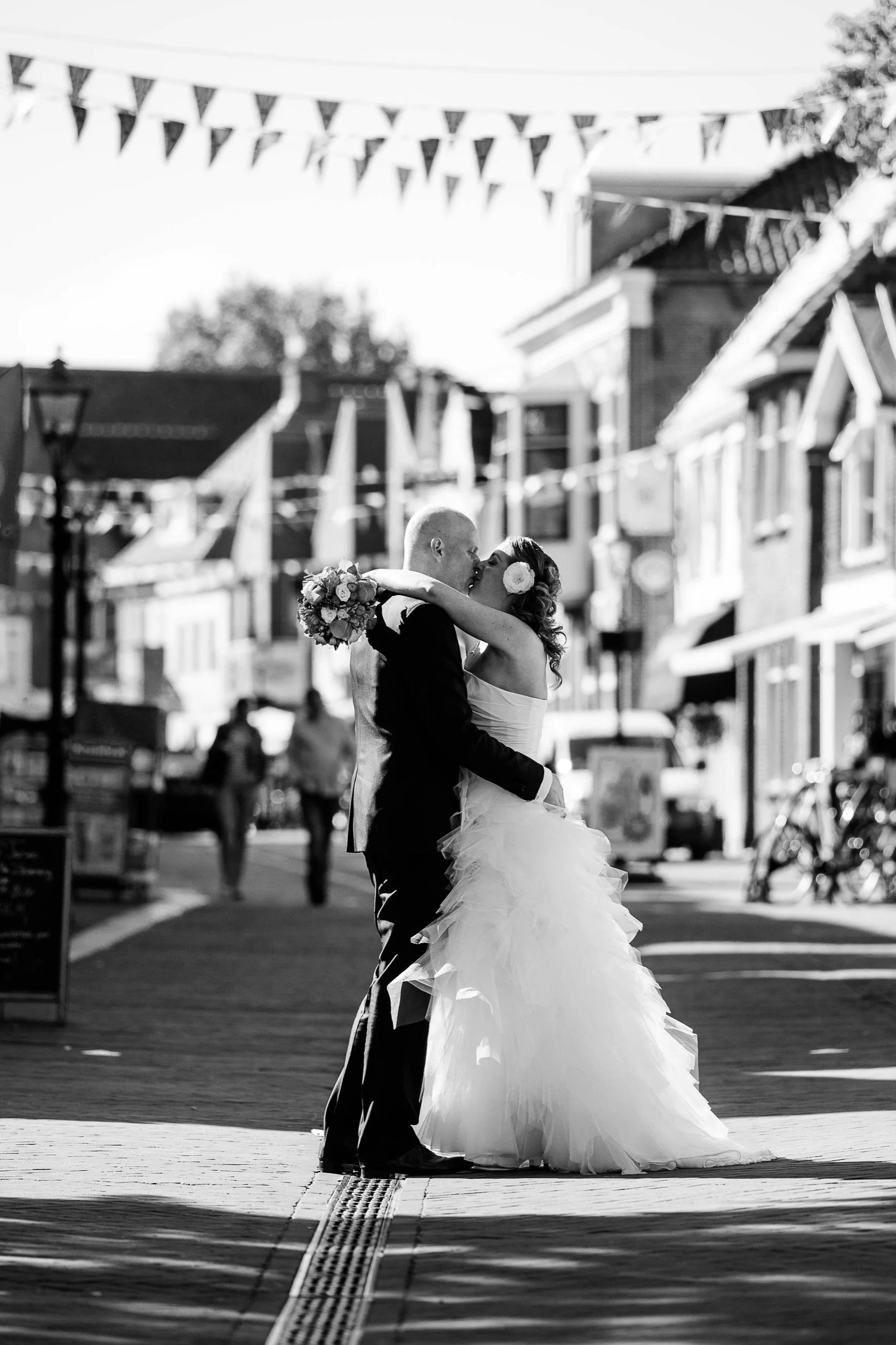 Bruidegom en bruid kussen op straat, omringd door winkelstraten en vlaggen, zwart-wil foto