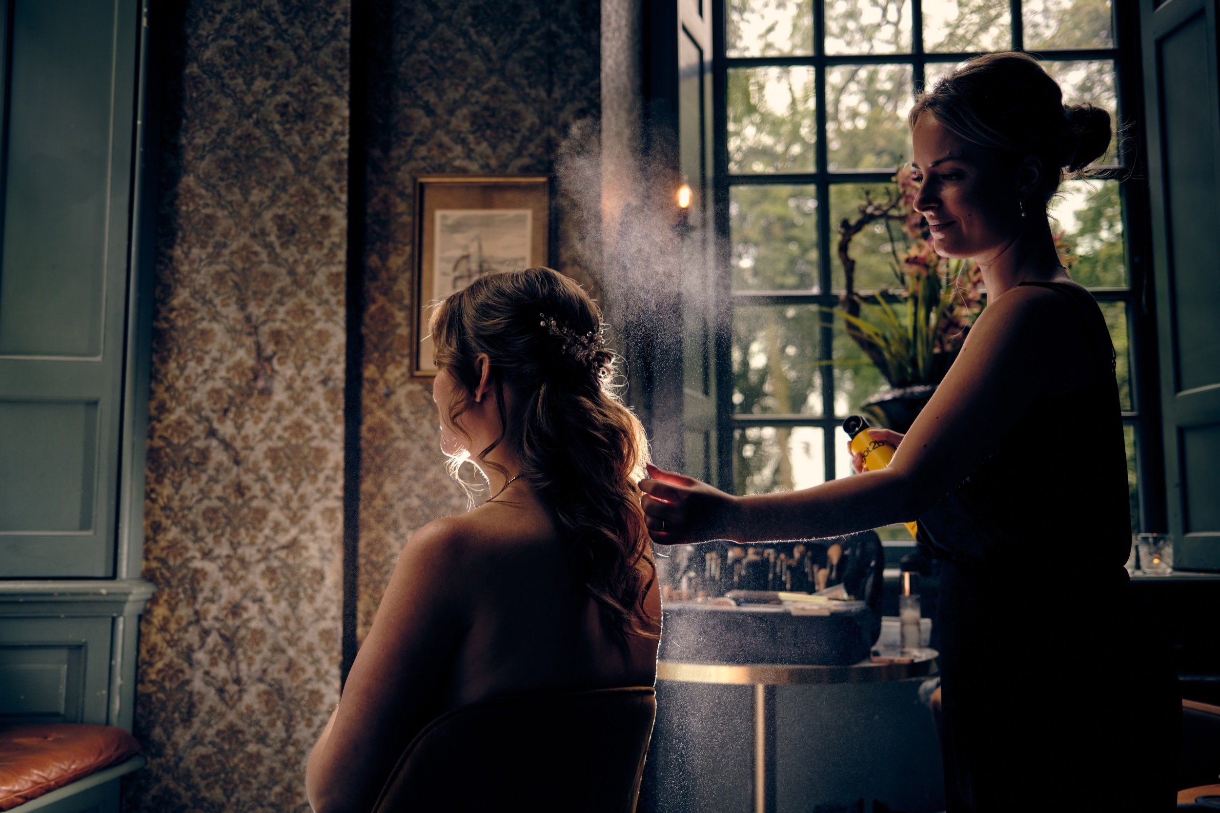 Vrouw die haar haar laat stylen door een stylist, die haar haar sprayt in een zonnig interieur met grote ramen en planten.